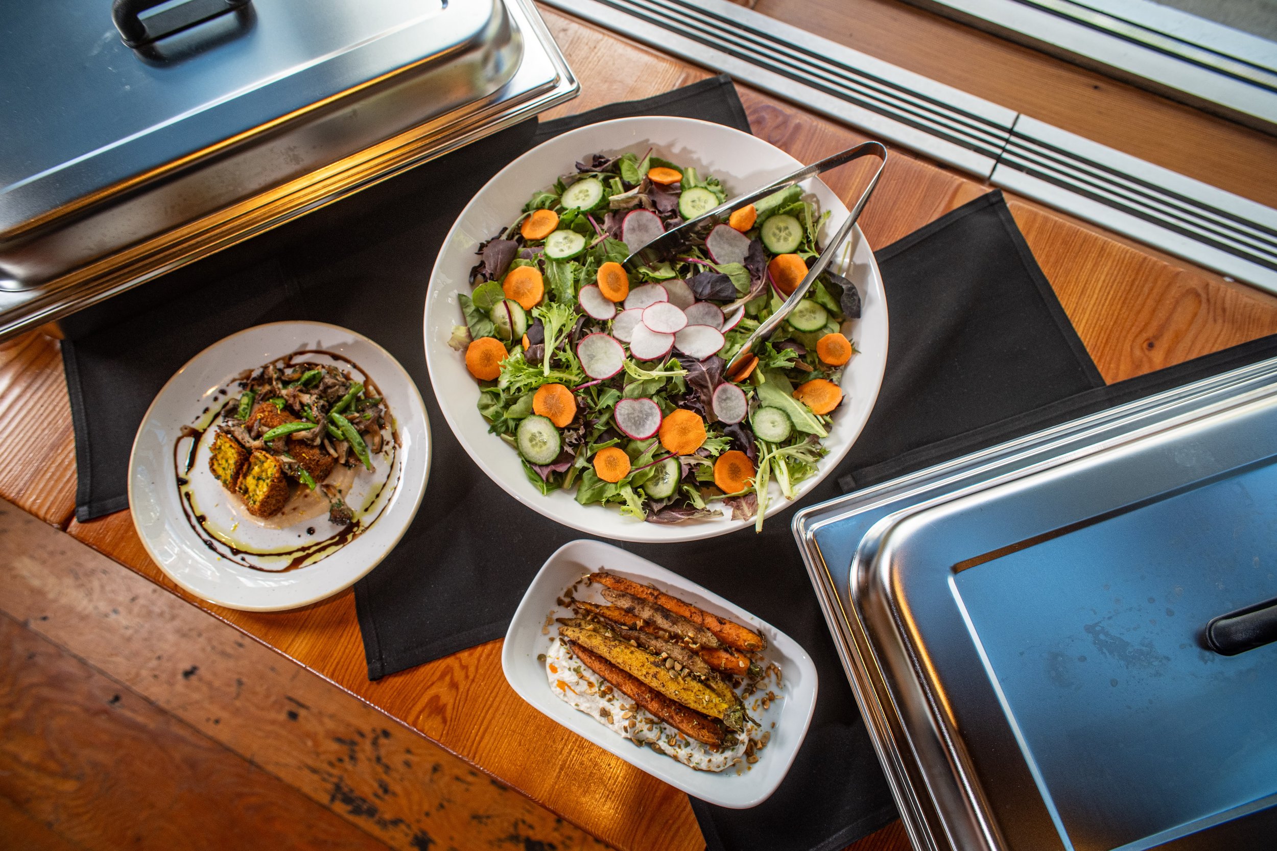 A buffet table with various dishes including a large green salad with cucumber, radish, and carrot slices, grilled vegetables, and other side dishes on white plates, set on a wooden table with a black tablecloth.