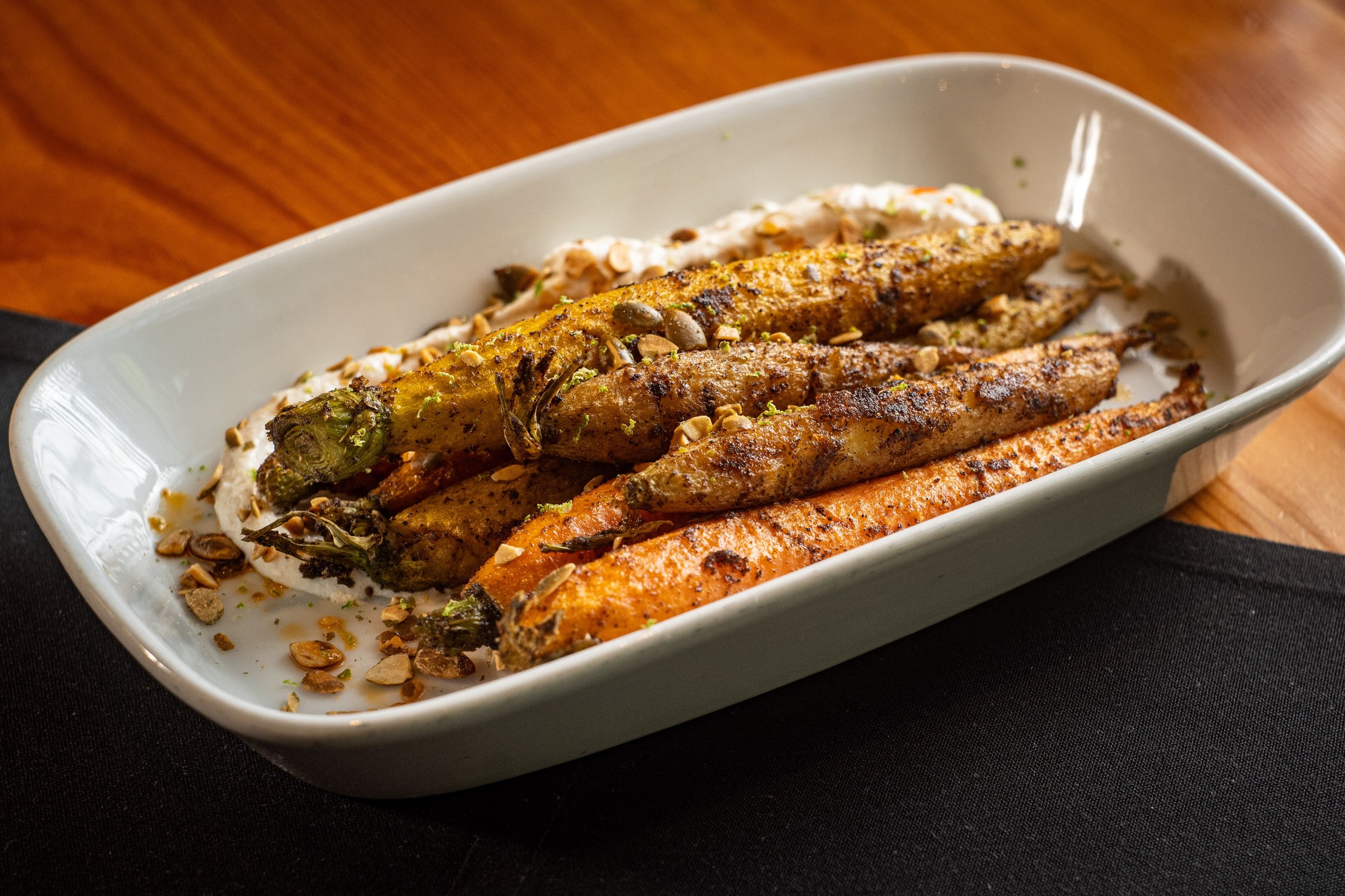 A white oval dish with grilled vegetables, including carrots, zucchini, and spring onion, topped with chopped nuts and a dollop of creamy sauce on a black table.