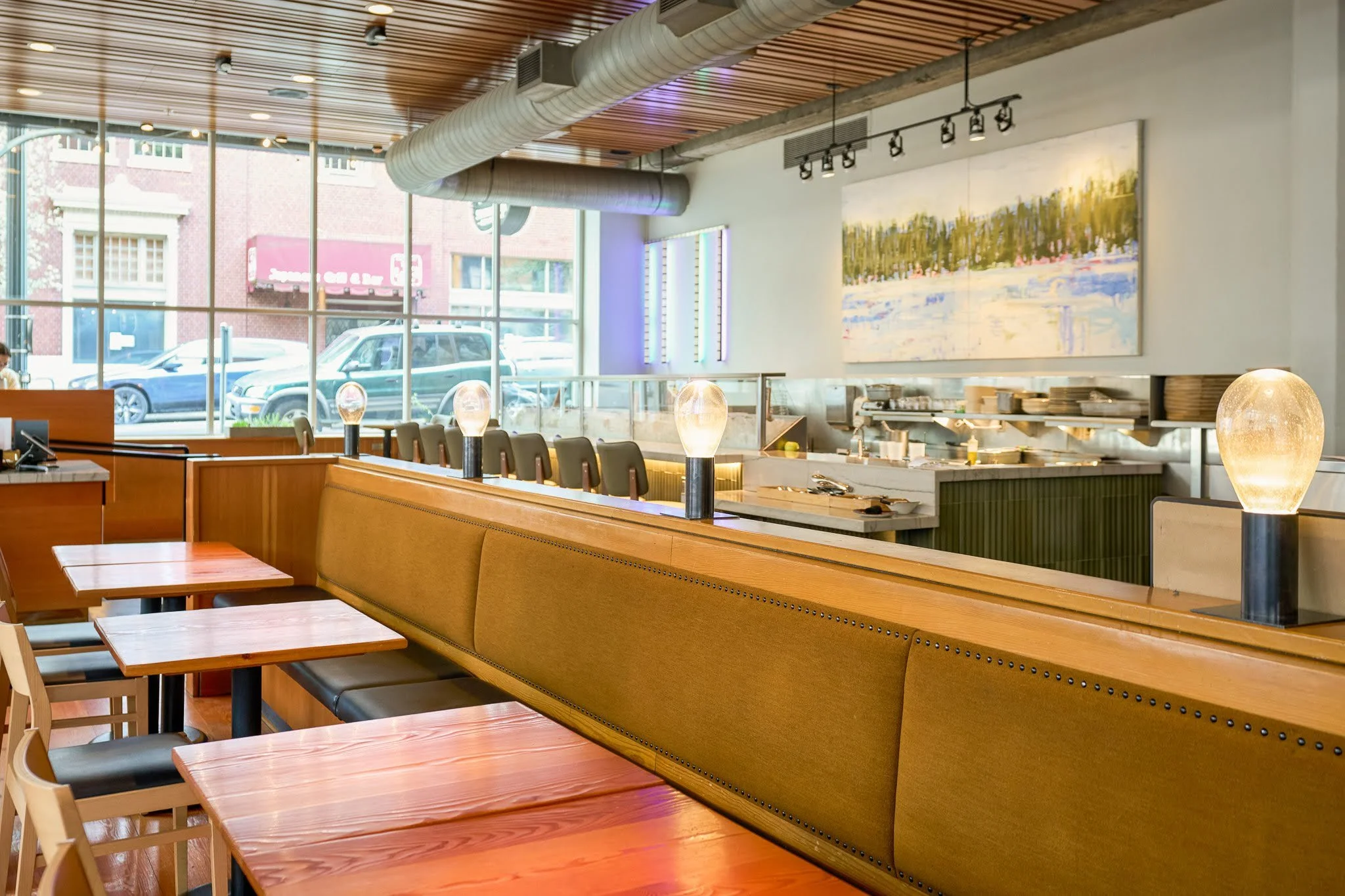 Interior of a modern restaurant with wooden tables and chairs, large windows, artwork on the wall, and decorative lighting fixtures.