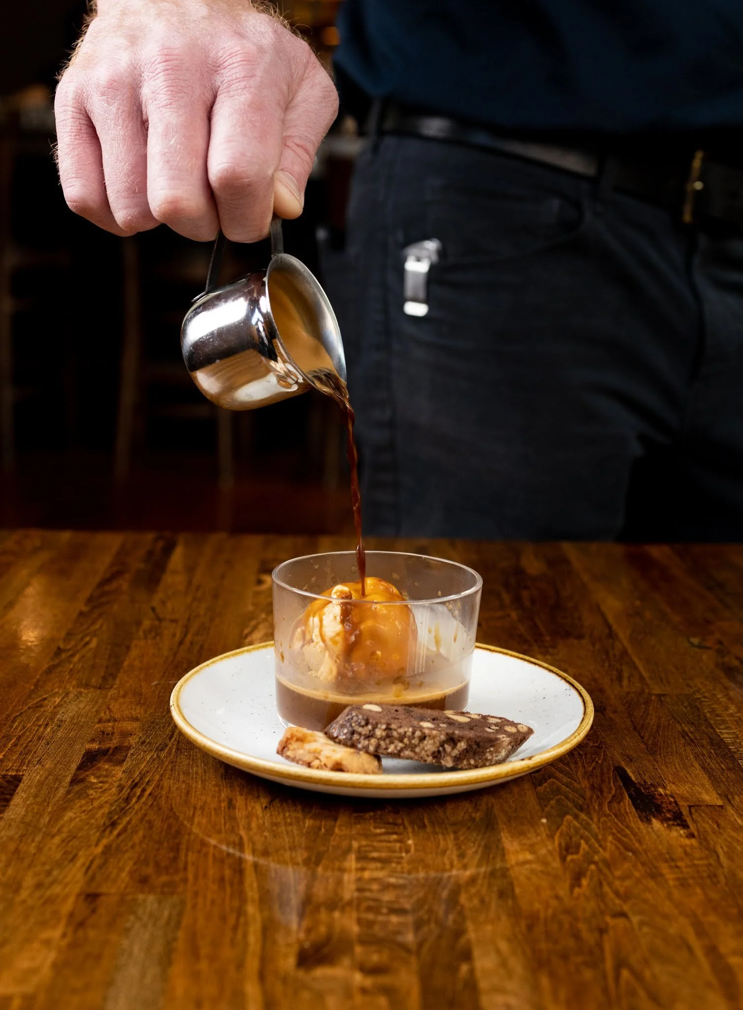 A person pouring hot coffee into a glass containing a scoop of vanilla ice cream, with assorted cookies on a plate in front, all on a wooden table.