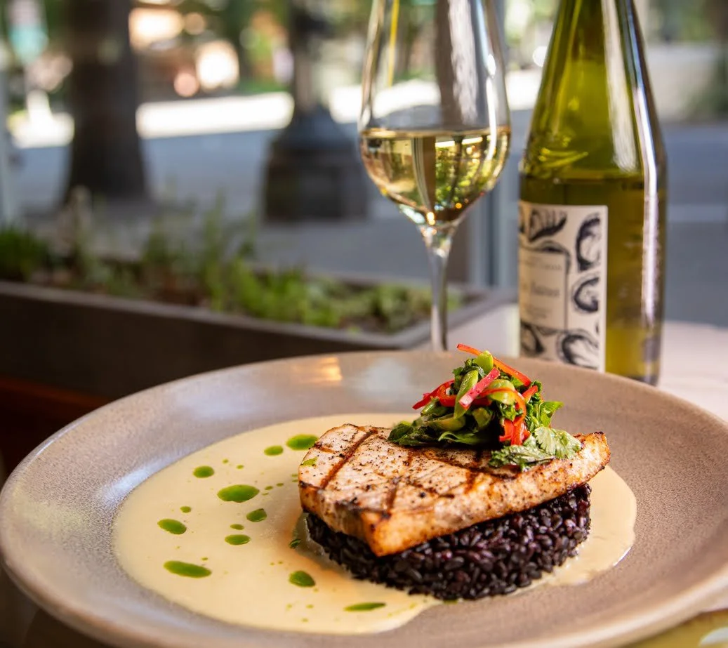 Grilled fish fillet served on a bed of black rice, topped with a small side salad, with a white creamy sauce and green oil drizzle around the plate. In the background, a glass of white wine and a bottle are visible, with a blurred outdoor view.