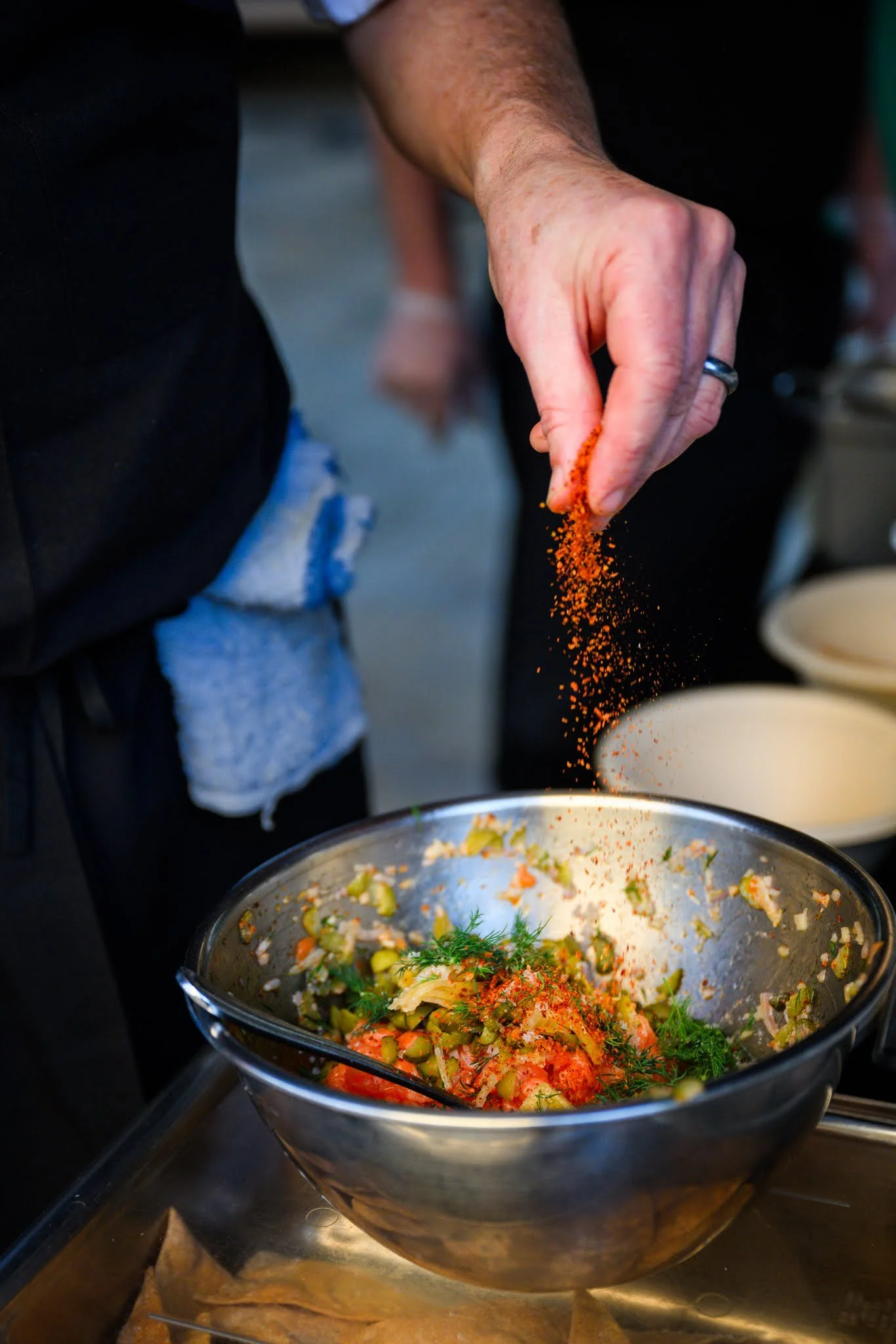 A person sprinkling a reddish-orange spice over a large mixing bowl containing chopped vegetables and herbs.