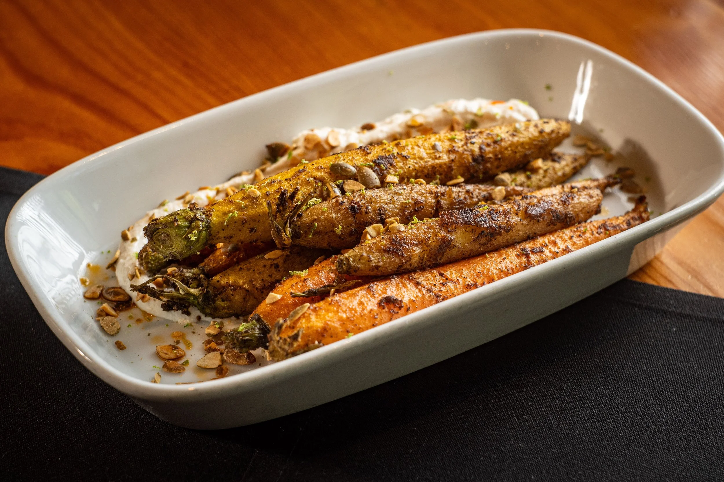 A white rectangular dish contains grilled vegetables, including carrots, and zucchini, topped with chopped nuts and served with a creamy dip or sauce garnished with herbs.