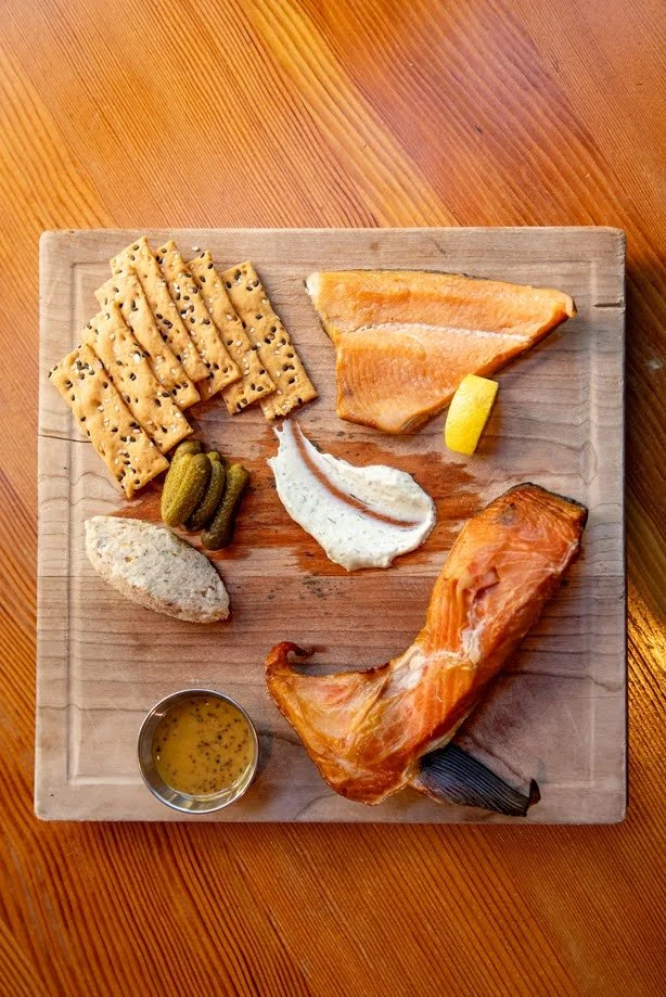 A wooden board with smoked salmon, cream cheese, crackers, pickles, lemon, and a cup of mustard seed honey.