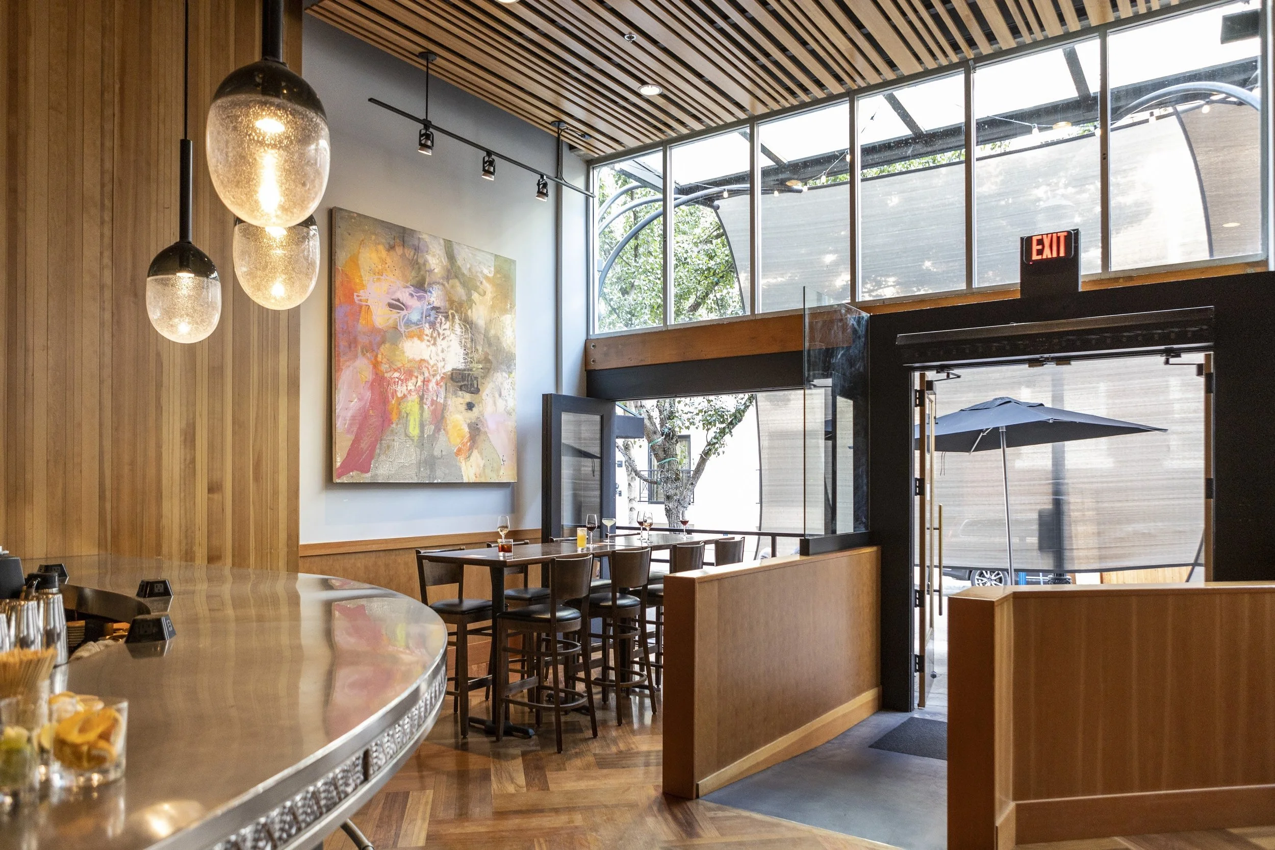 Interior of a modern restaurant with wooden wall paneling, large abstract painting, pendant lights, tall glass windows, and outdoor patio area with umbrella.