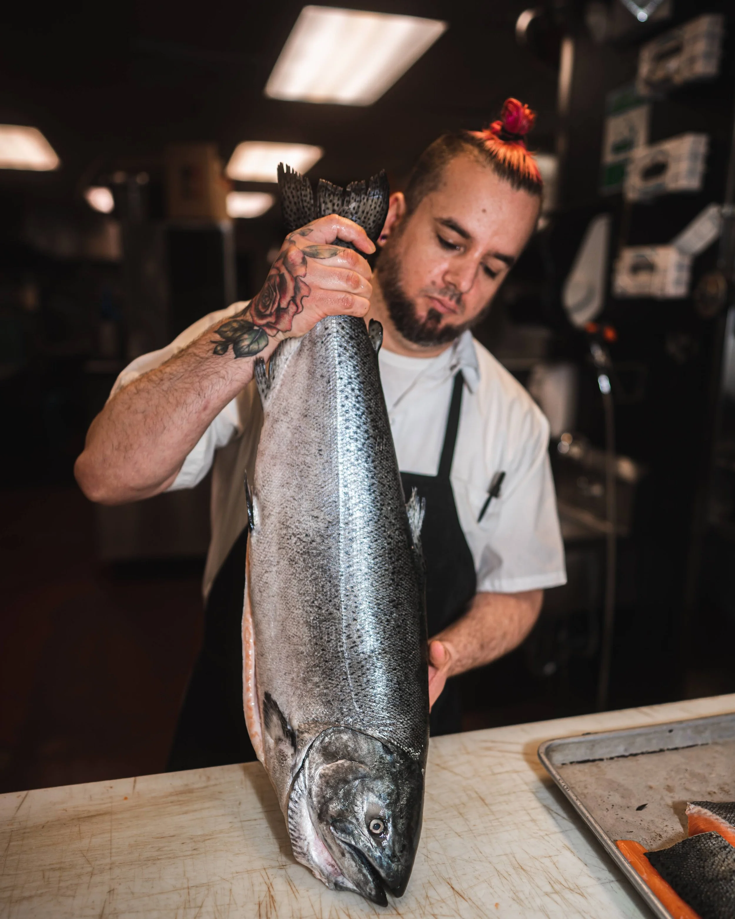 A person with tattoos on their arm holding a large freshwater fish by the tail, with a cutting board and fish fillet on tray nearby, in a kitchen setting.