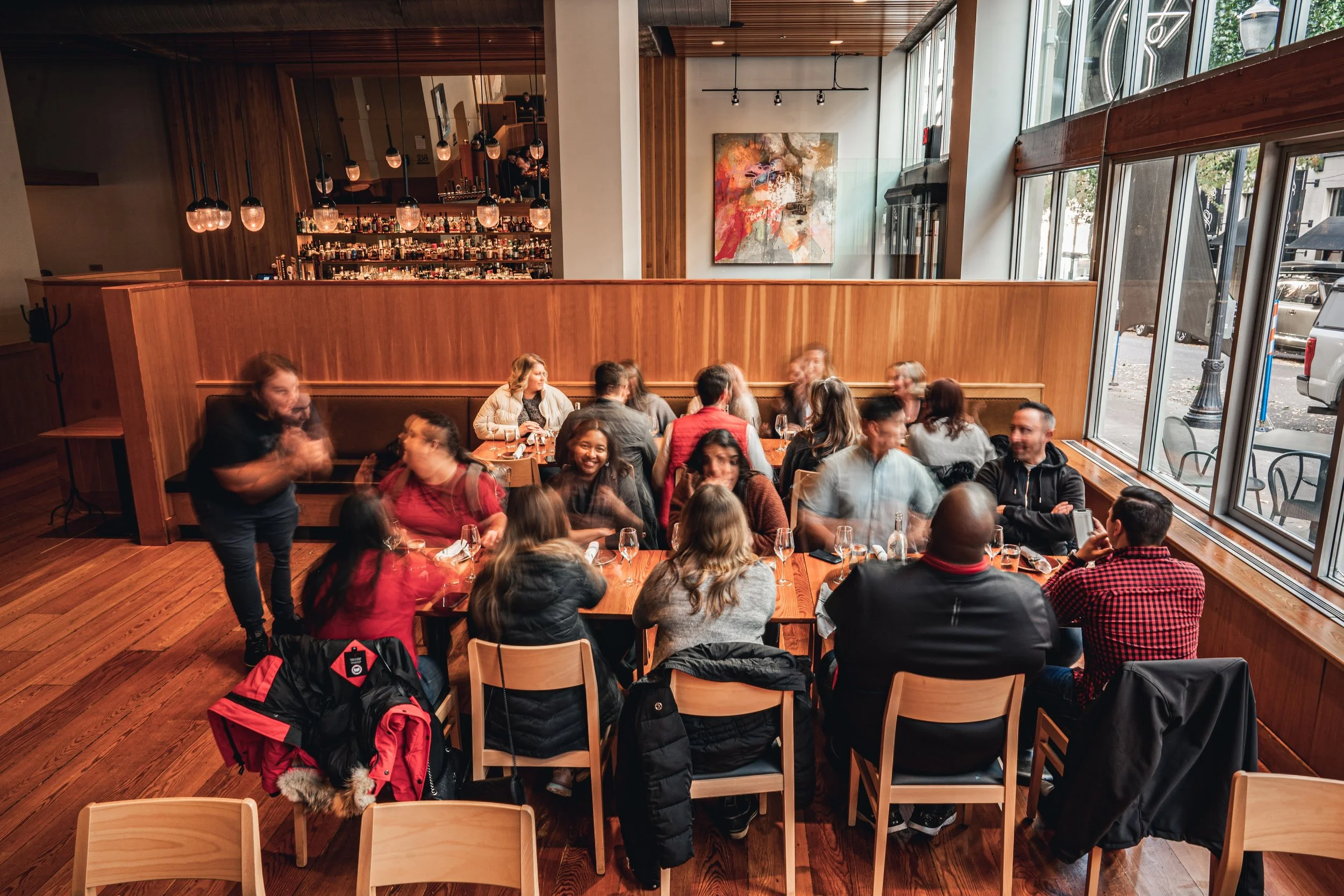 A lively restaurant interior filled with people sitting at wooden tables, enjoying their meals and conversations. The restaurant has large windows allowing natural light, a wooden decor, and an abstract painting on the wall. The bar area with bottles