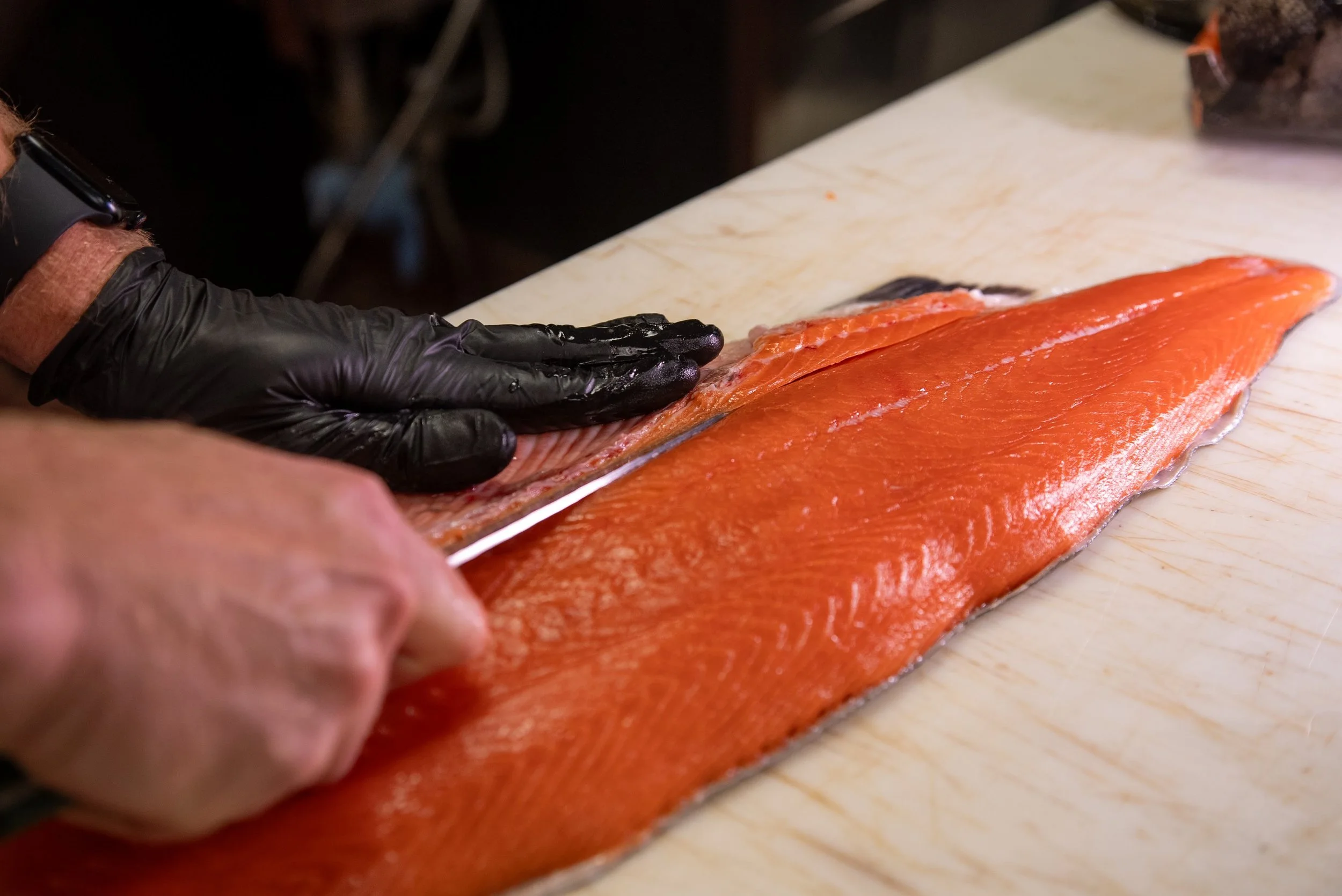 A person wearing black gloves slicing a large piece of fresh, orange salmon on a cutting board.