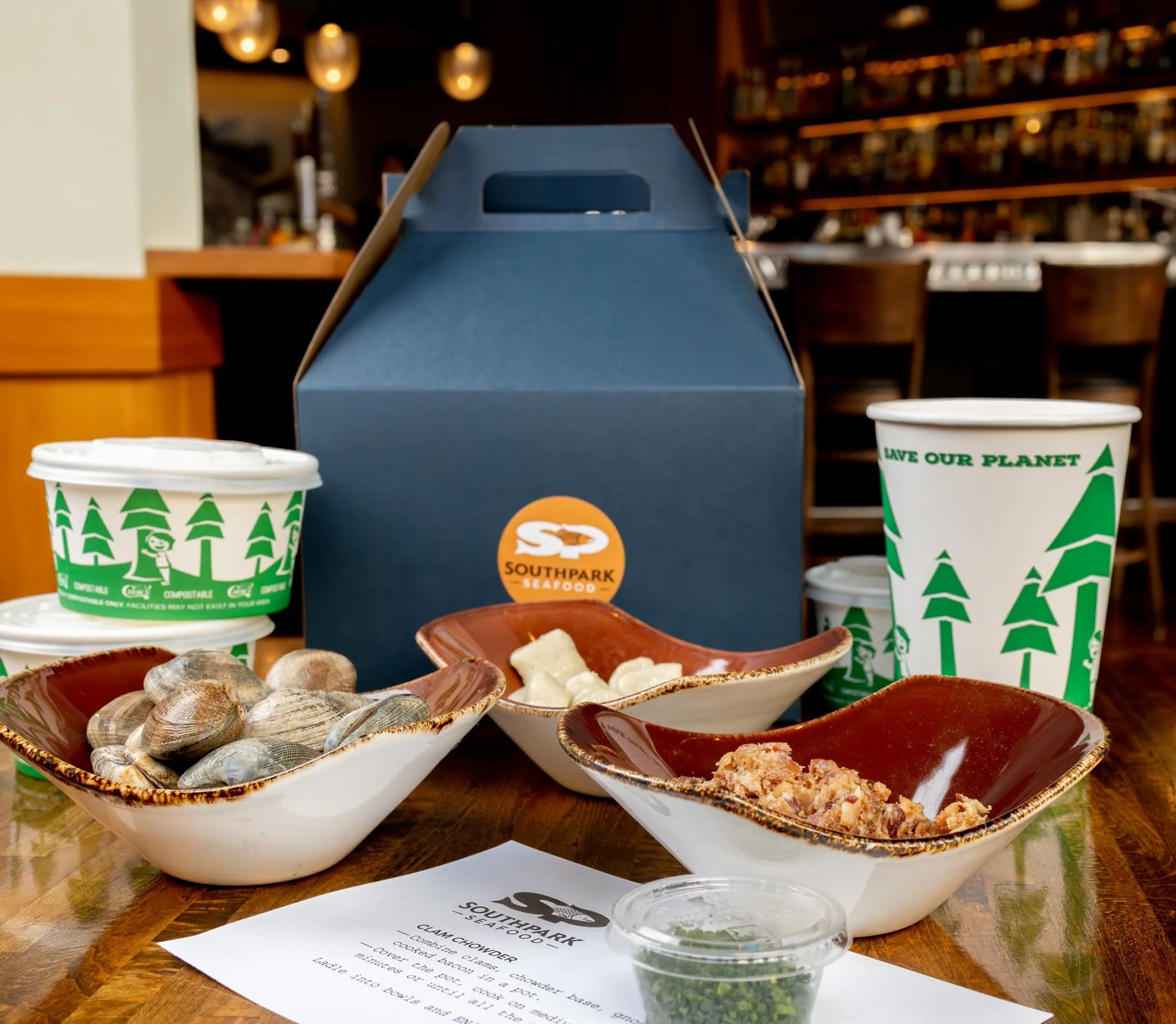 Several bowls of seafood, including clams and oysters, on a wooden table at Southpark Seafood restaurant, with a blue takeout box and paper menu in the background.
