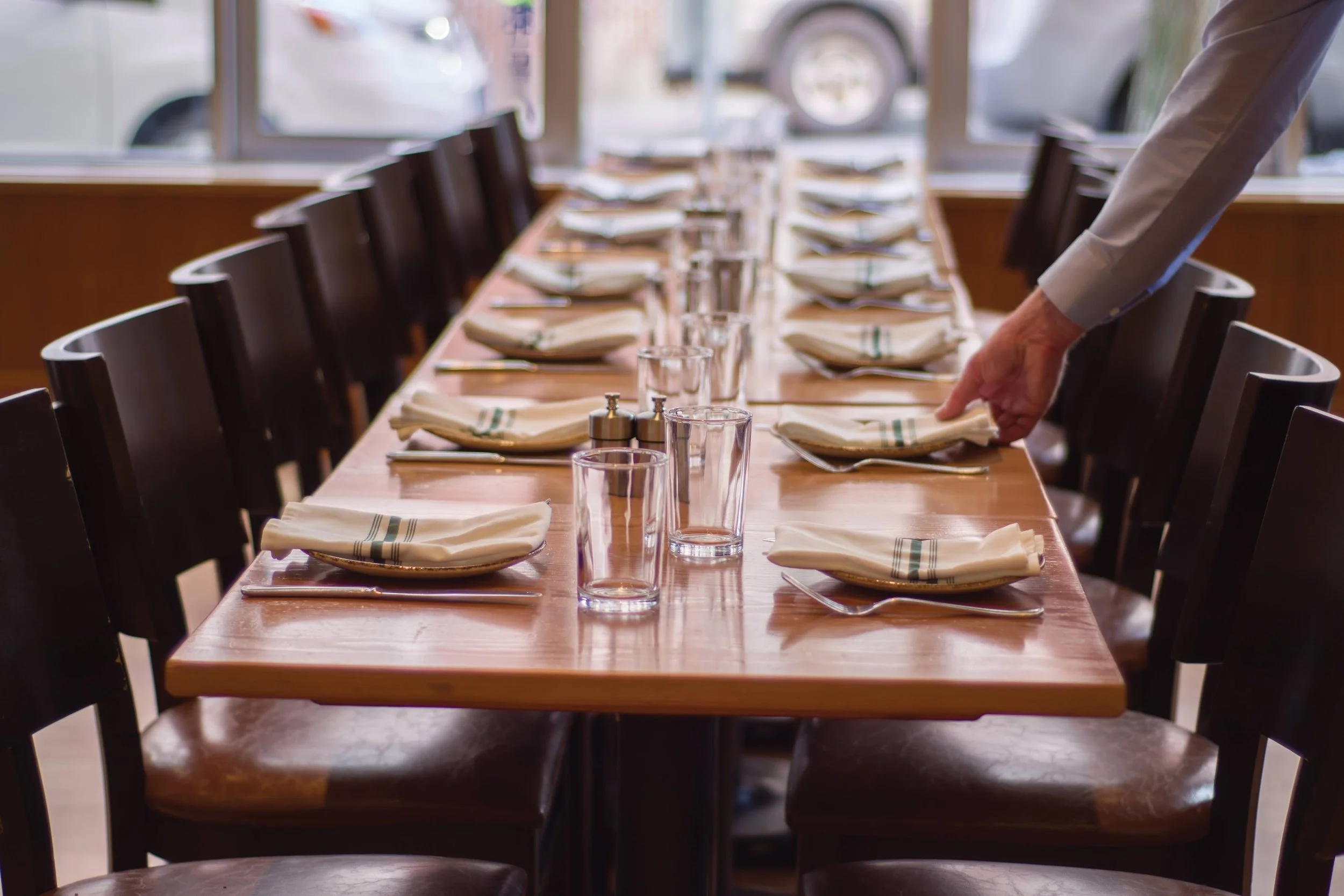 A long wooden restaurant table is set with white napkins, transparent glasses, silverware, and small salt and pepper shakers, ready for diners. A person's arm is arranging the table setting. Large windows in the background reveal parked cars outside.