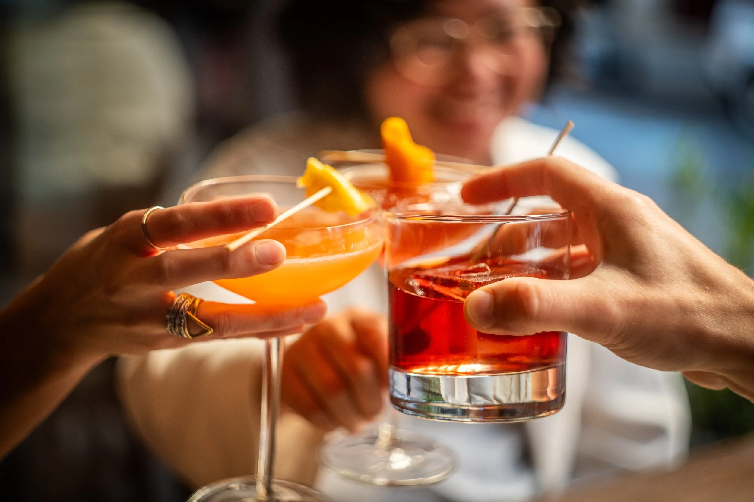 Two women toasting with colorful cocktails in a social setting, one in an orange drink and the other in a red drink, both garnished with fruit slices.