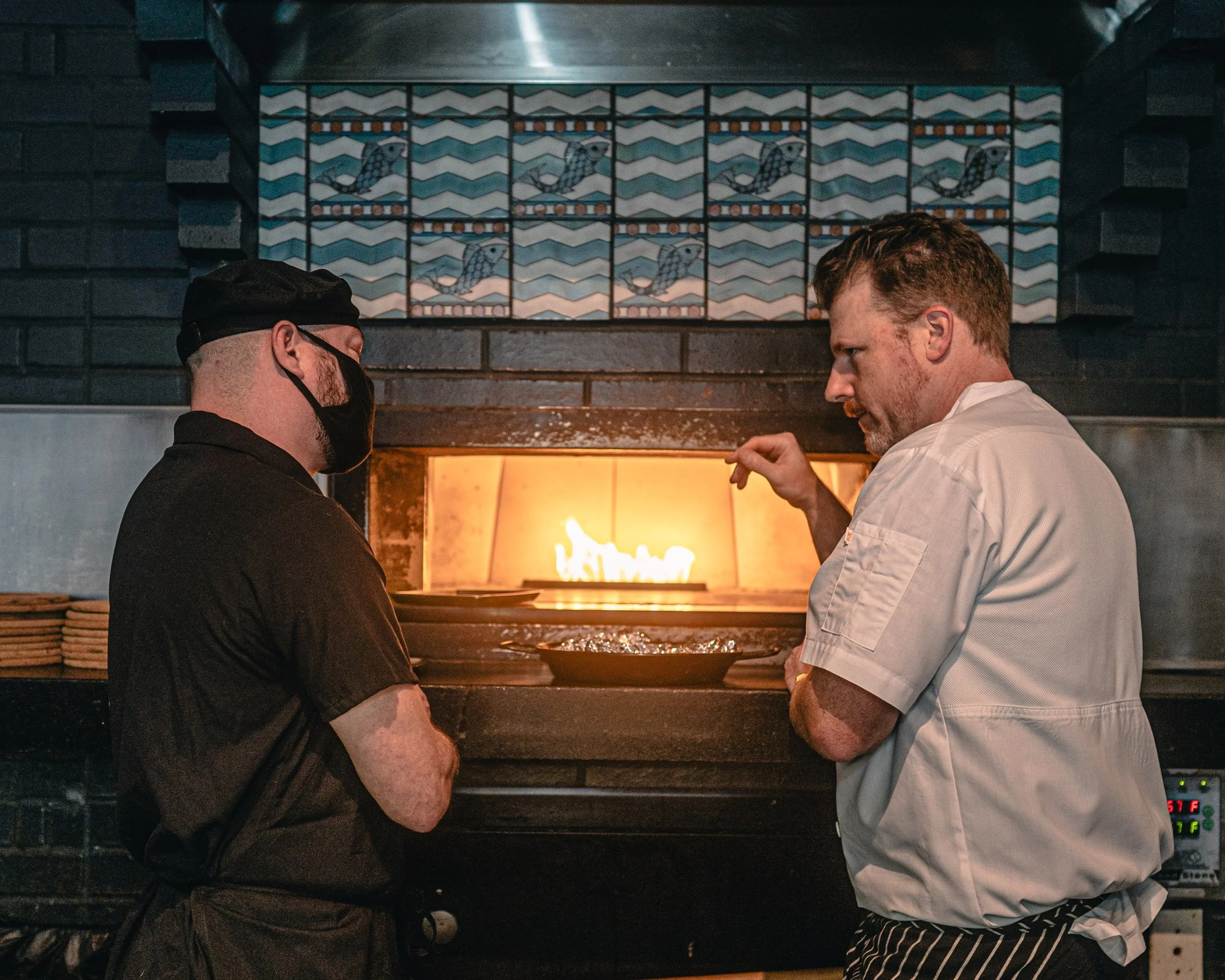 Two chefs stand in front of a pizza oven, one wearing black and a face mask, the other in a white shirt, discussing a pizza with flames inside the oven. There are decorative tiles with a bird pattern above them.