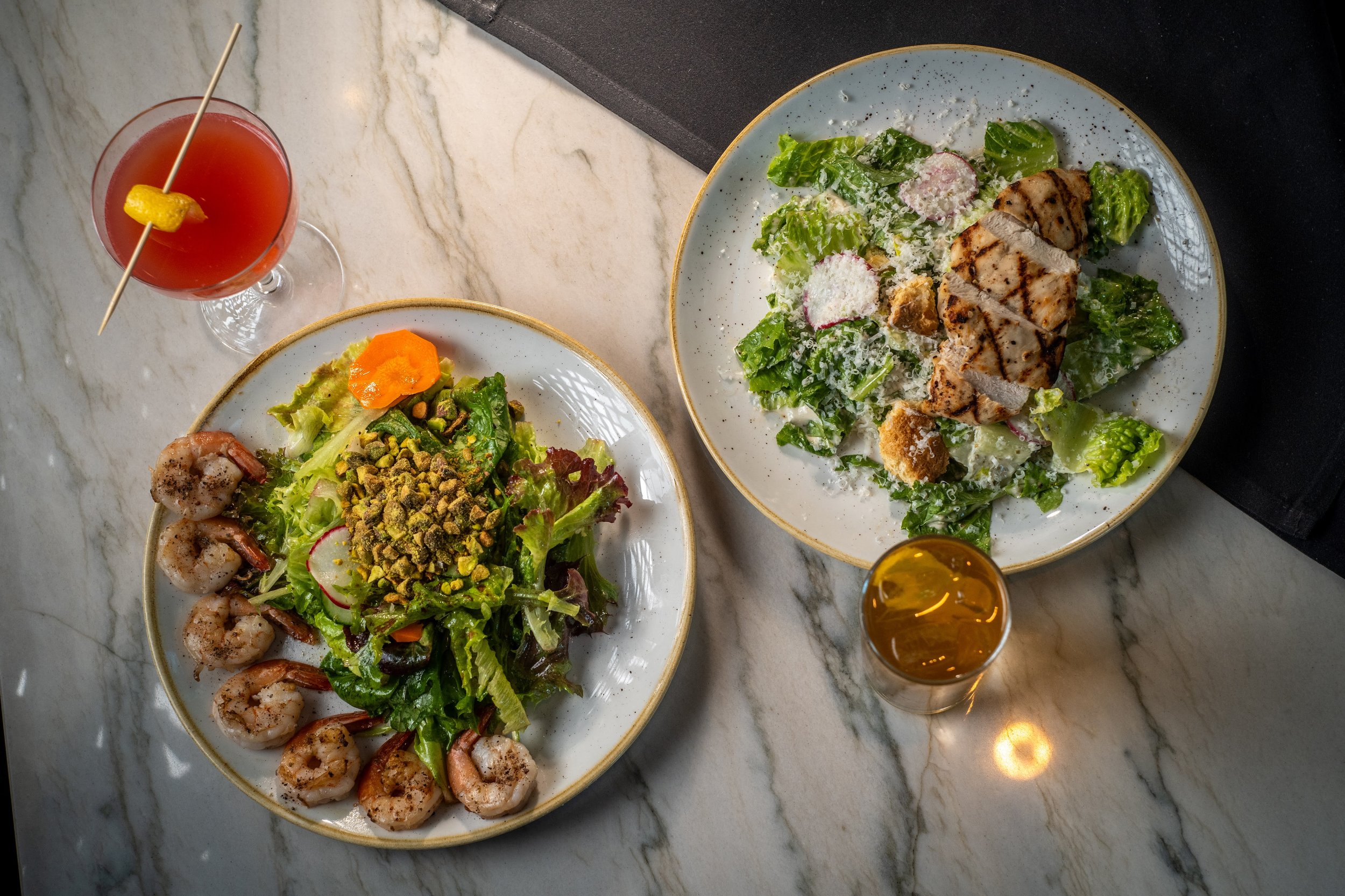 Two plates of salad, one with grilled chicken, lettuce, radishes, and Parmesan cheese, and the other with shrimp, mixed greens, radishes, pistachios, and a yellow dressing, on a marble table. Two beverages are also present, one pink drink garnished w