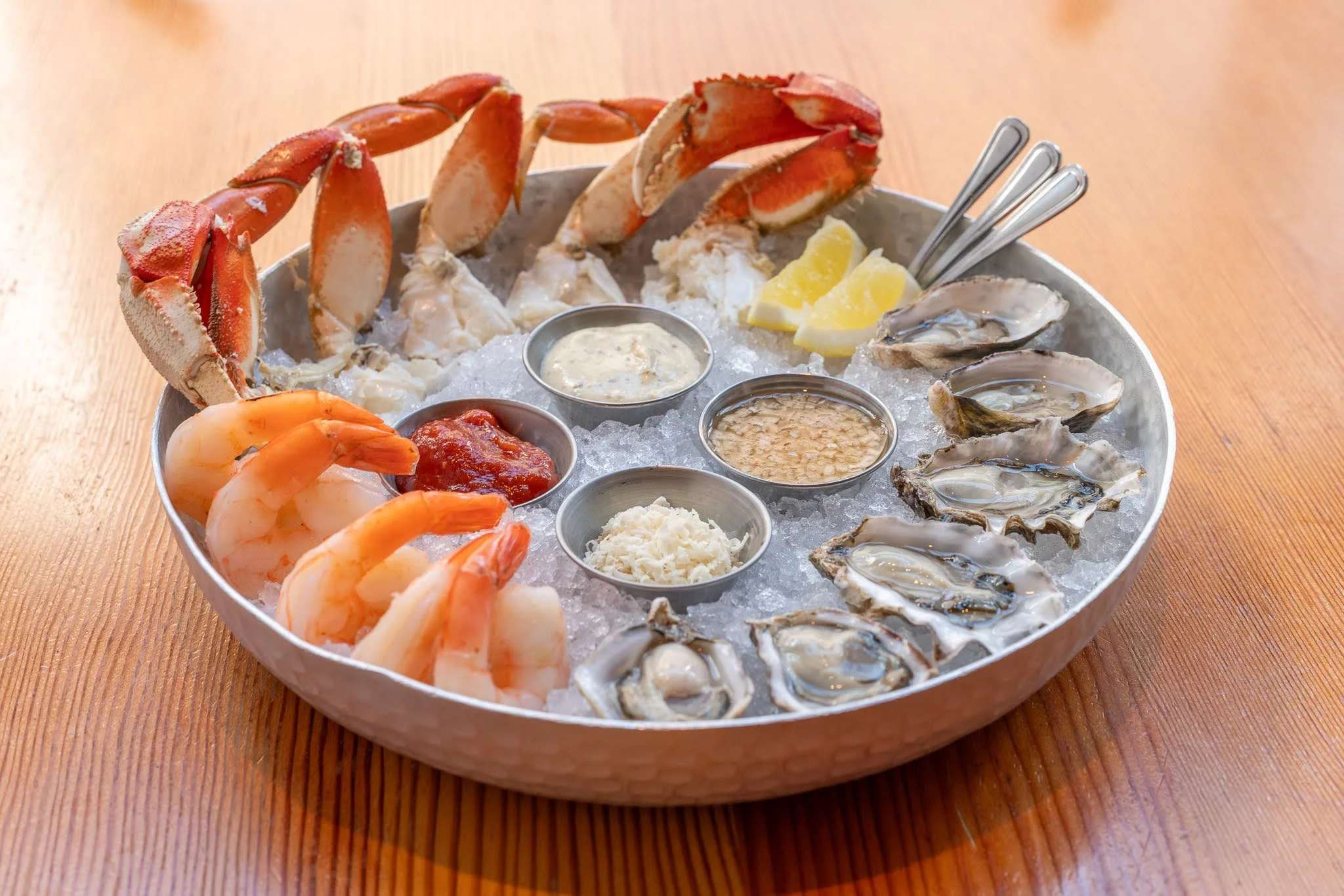 A round seafood platter with crab legs, shrimp, oysters, lemon wedges, and various dipping sauces on a bed of crushed ice, placed on a wooden table.