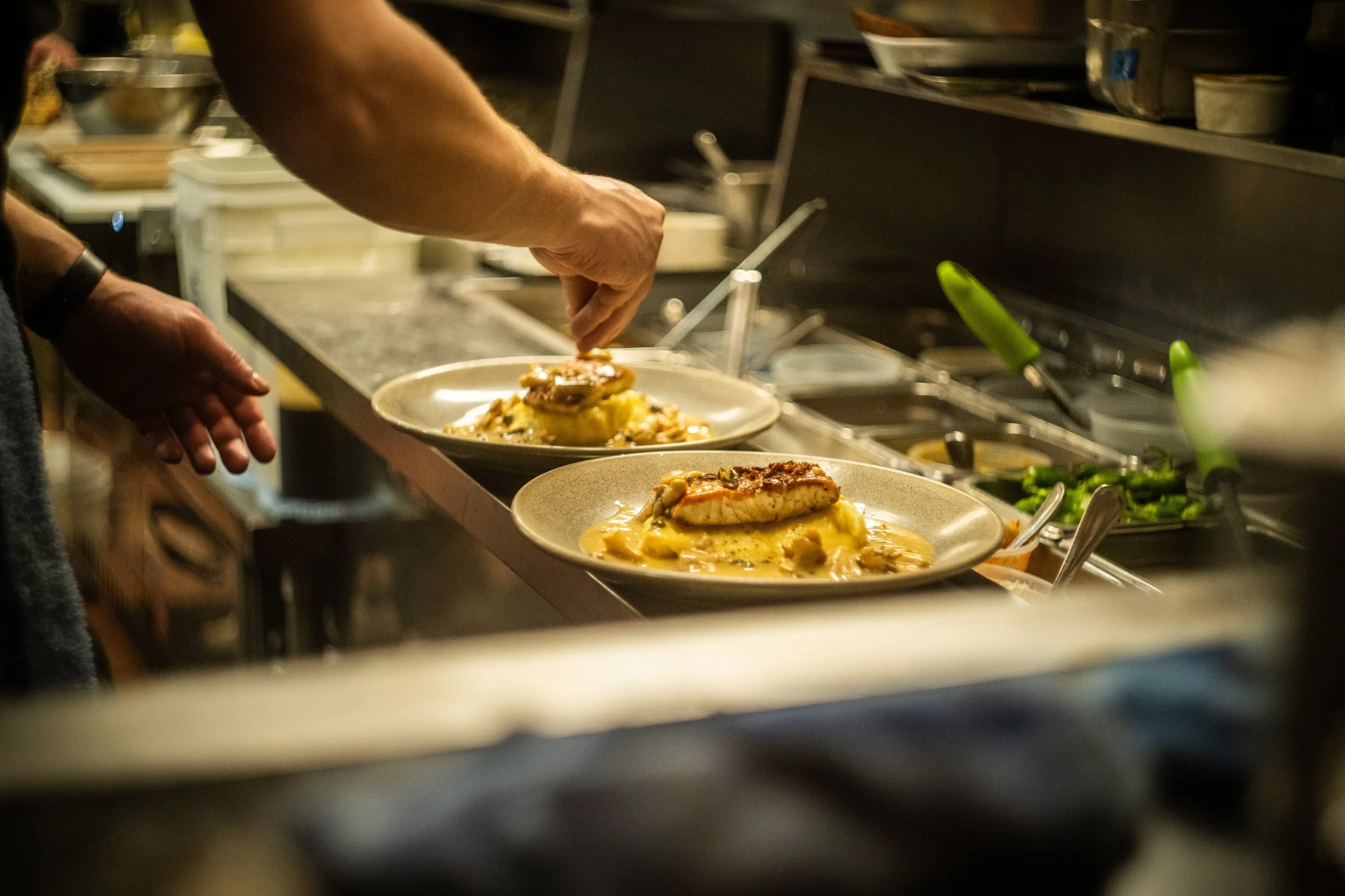A chef or cook preparing dishes in a professional kitchen, with plated meals on a counter and various kitchen tools and ingredients in the background.