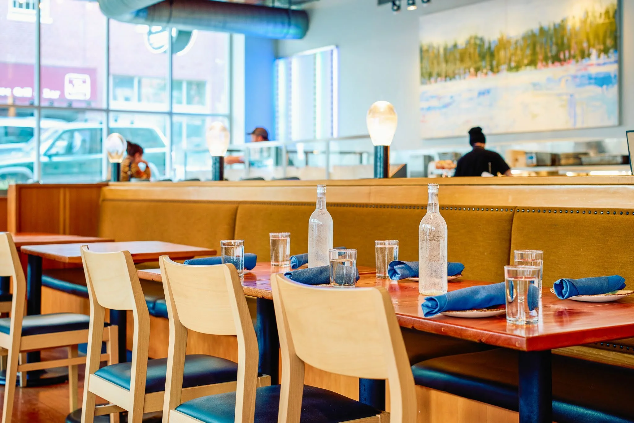 Interior of a modern restaurant with wooden tables set with glasses and blue cloth napkins, light wood chairs, large windows letting in natural light, and a mural on the wall in the background.