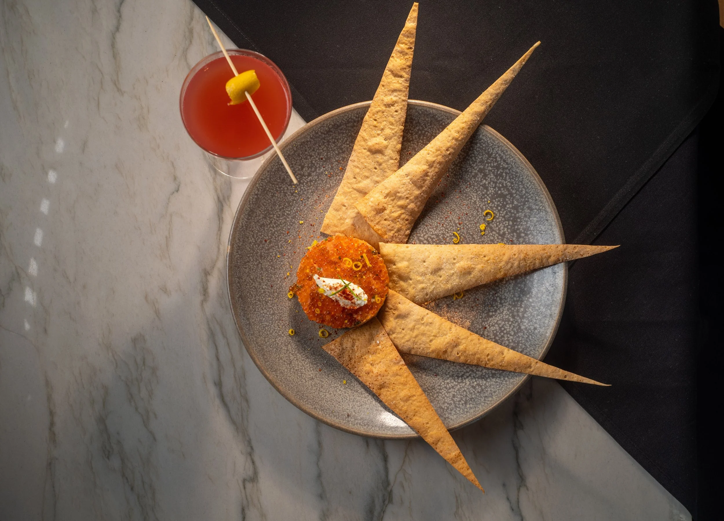 A round grey plate with four triangular baked tortilla chips arranged around a small serving of red caviar with a dollop of cream and yellow garnish on top. A cocktail glass with red drink and a lemon slice garnish is placed nearby on a marble surfac