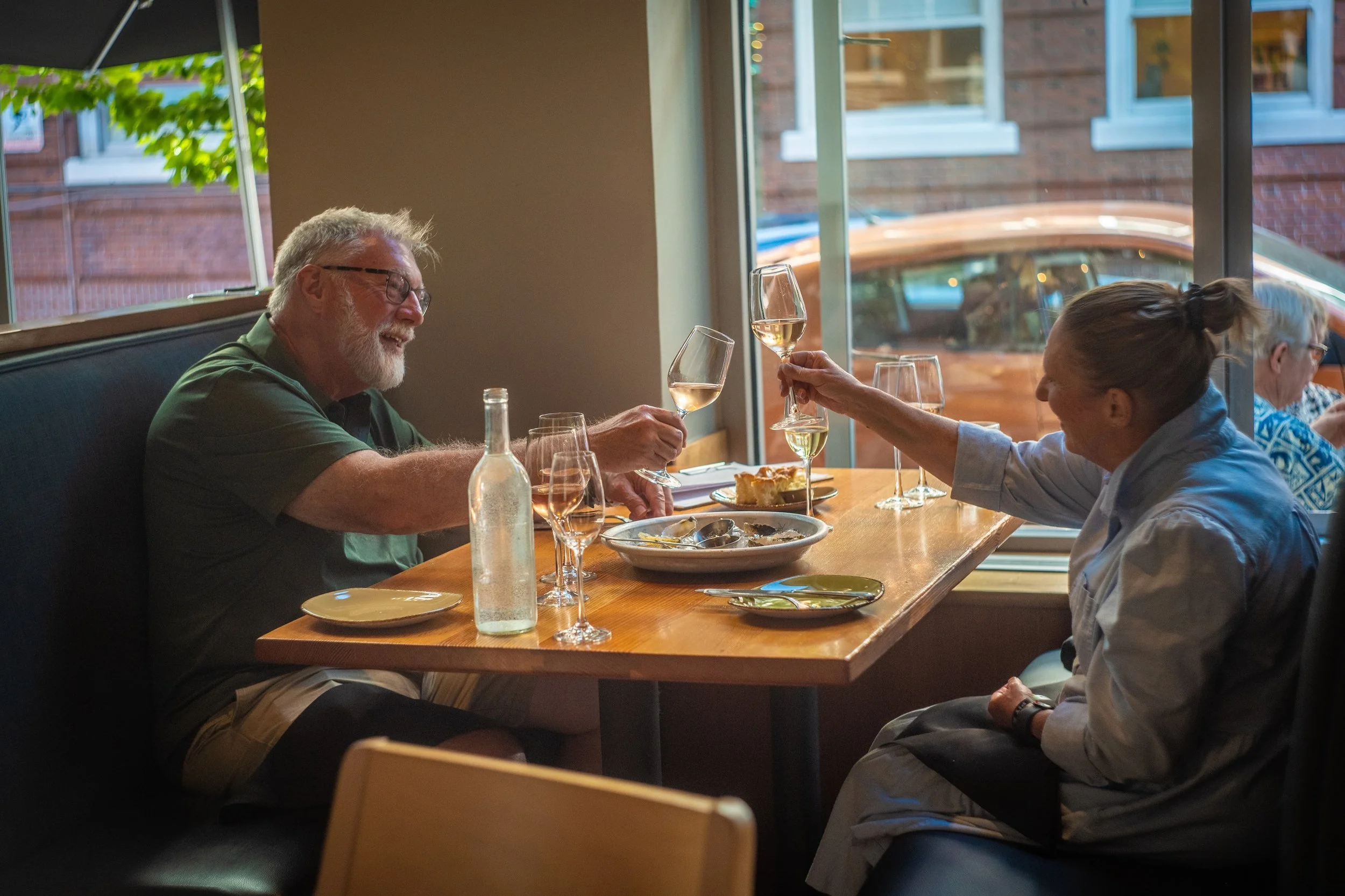 Older man with glasses and a beard, wearing a green shirt, smiling and clinking a glass of white wine with a woman with braided hair, wearing a light jacket, in a restaurant with large windows, during daytime.