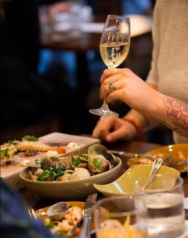 Person holding a glass of white wine at a dining table with seafood and dishes.