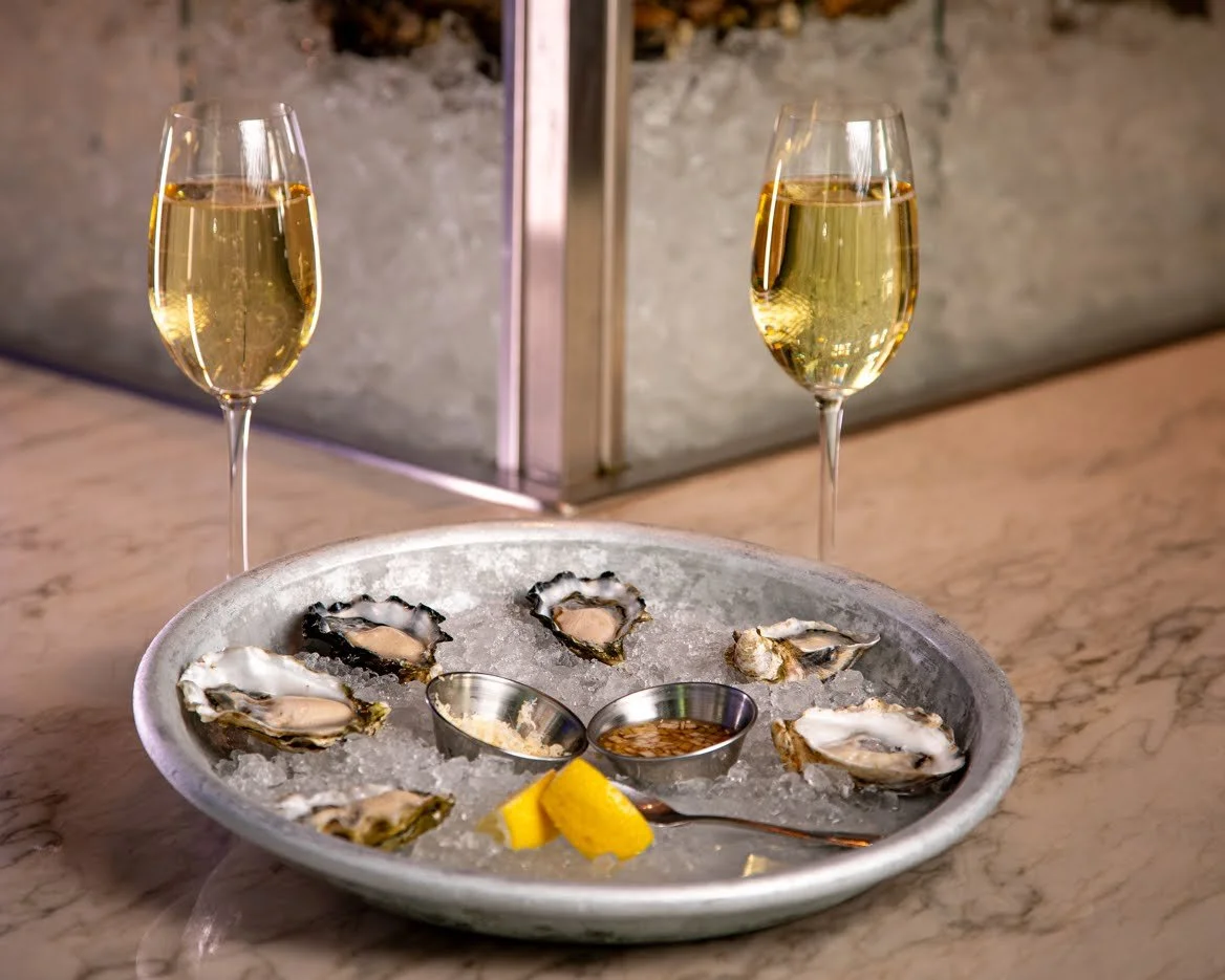 A plate of fresh oysters on ice with lemon wedges, two glasses of champagne, and small containers of dipping sauces on a marble countertop.