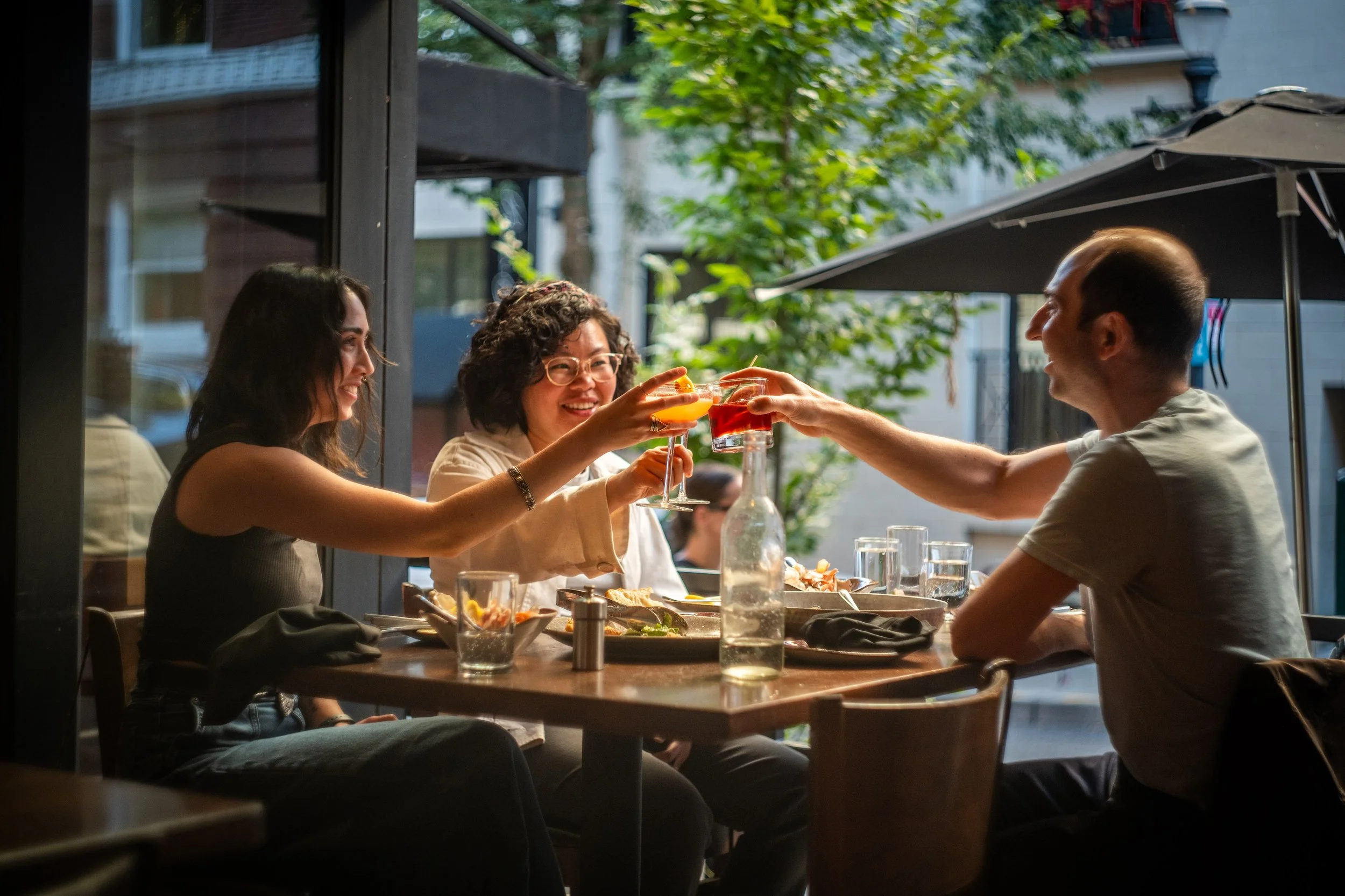 Four friends are sitting at a dinner table outside, raising their glasses filled with colorful drinks in a toast, smiling, with sunlight filtering through green leaves overhead.