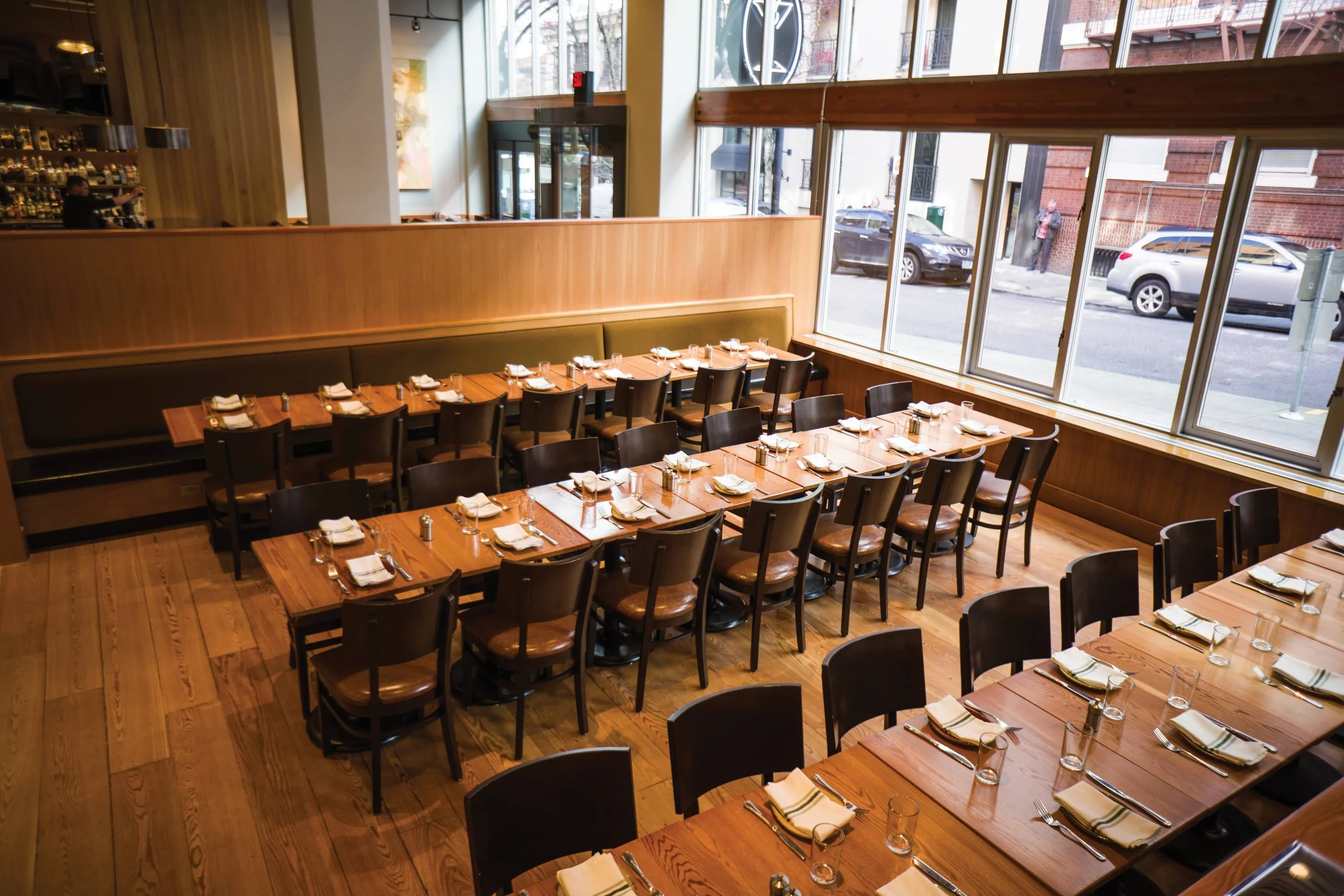A restaurant with wooden tables and chairs set with napkins, plates, silverware, and glasses. Large windows on one side of the room reveal parked cars and a busy street outside.