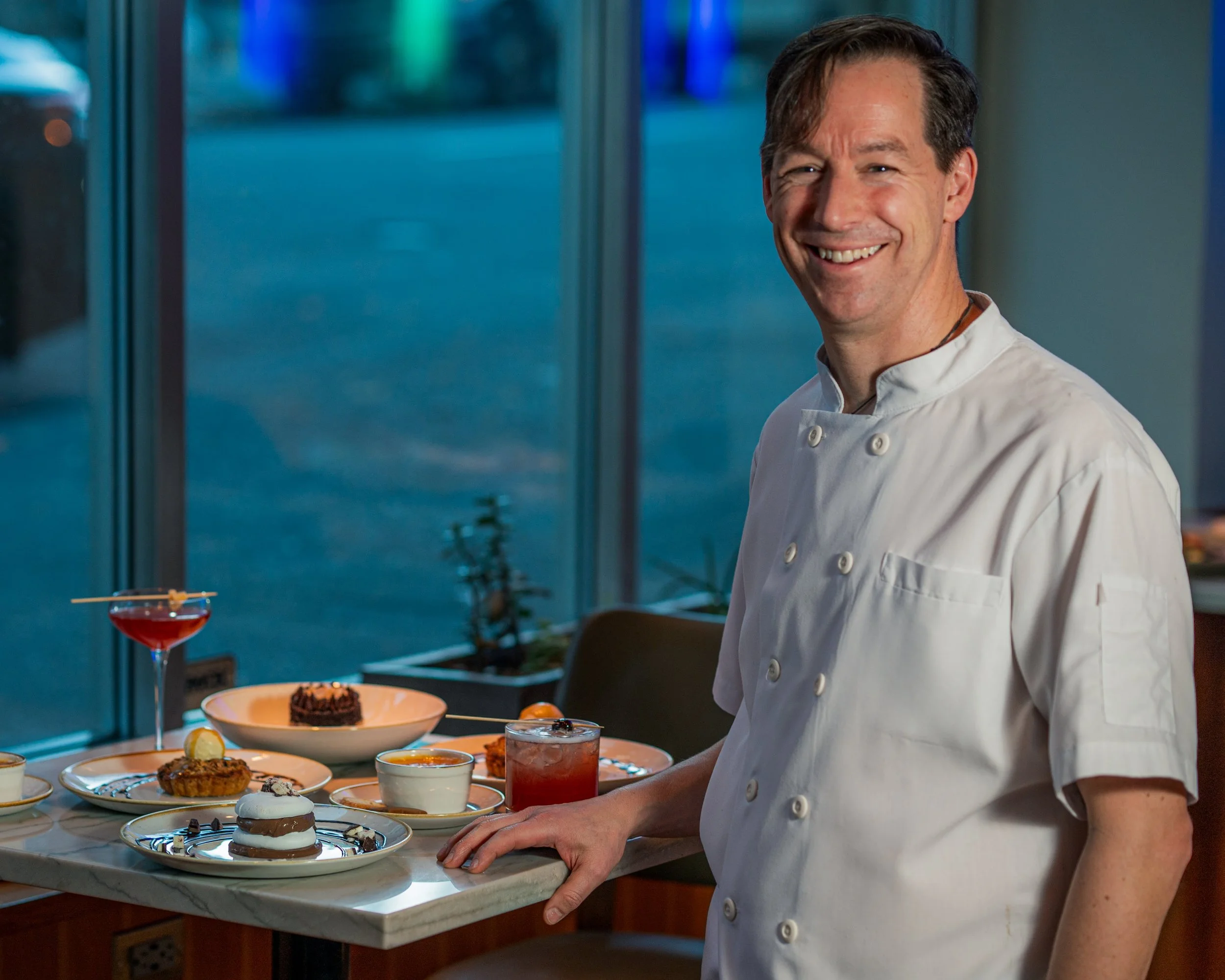 A male chef in a white uniform standing near a table with desserts, including a chocolate cake, layered pastry, and various drinks, in a restaurant setting with blue-tinted lighting.