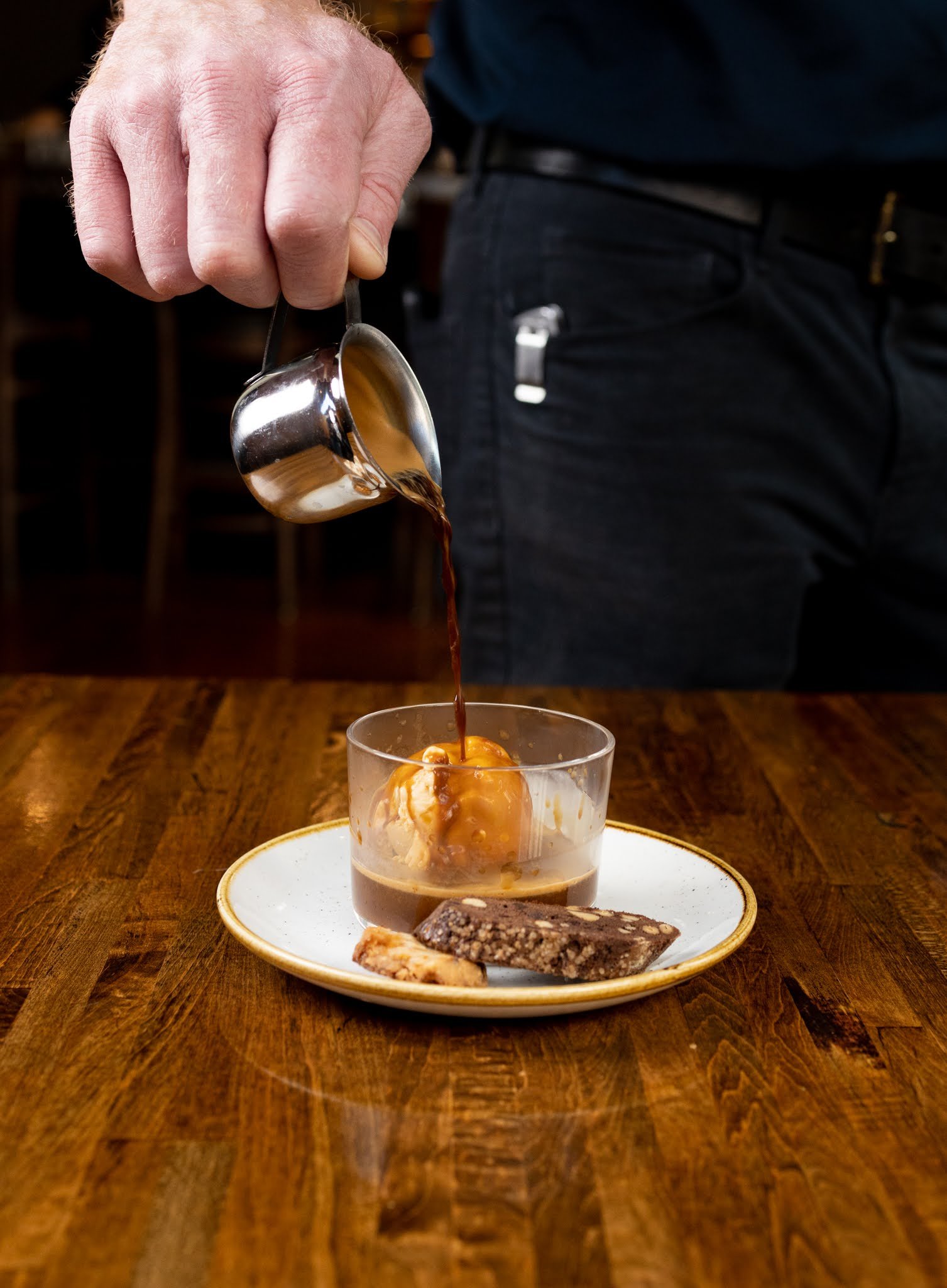 A person is pouring hot coffee into a glass with ice cream, on a wooden table with a plate of assorted cookies in front.