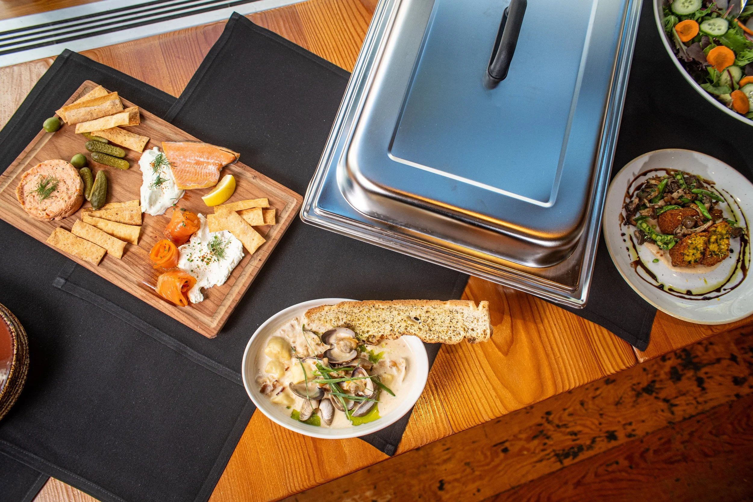 A wide variety of dishes including a cheese platter with smoked salmon, vegetables, a bowl of chowder with mussels and herbs, and a salad with cucumbers and carrots, on a wooden table with a chafing dish in the background.