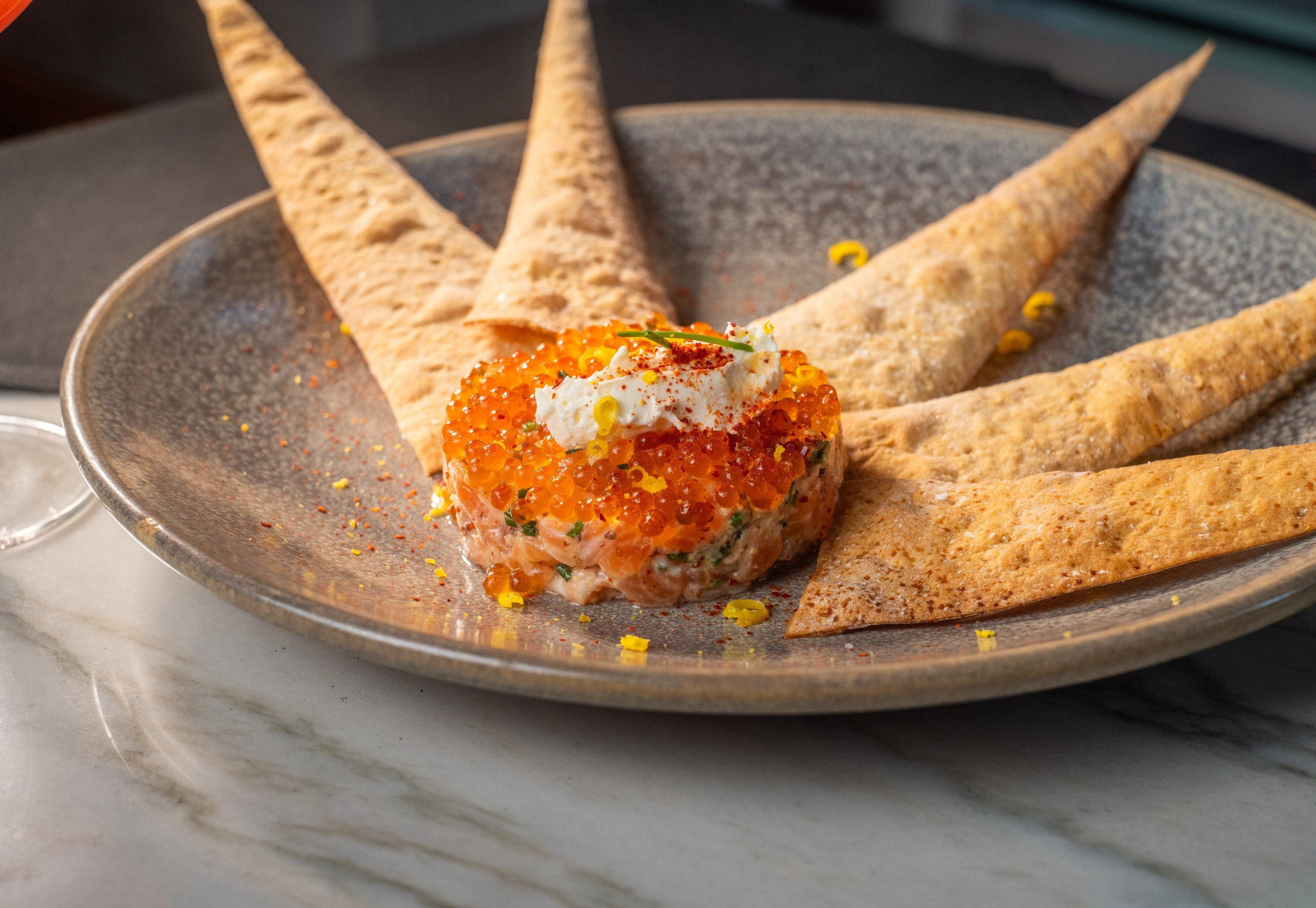 A seafood dish with salmon tartare topped with orange fish roe, garnished with herbs and a dollop of cream, served with crispy flatbread on a textured metal plate.