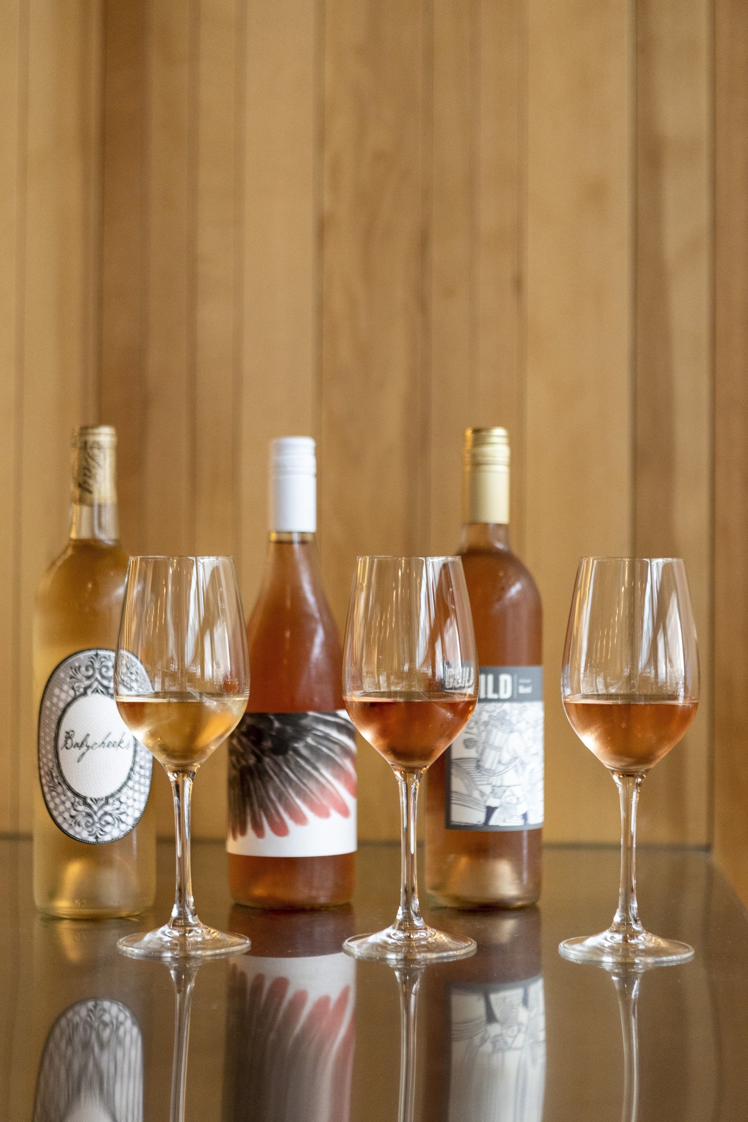 Several wine glasses filled with rosé wine and bottles of wine in the background against a wooden wall.