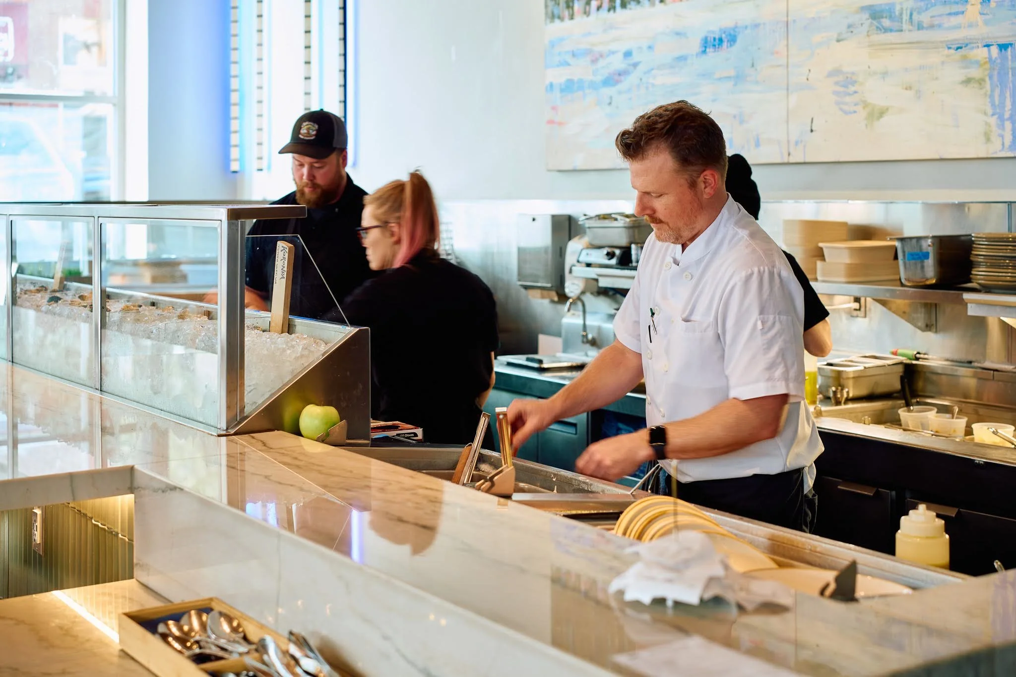 Employees working behind a food counter in a restaurant, with ice and utensils in the foreground and abstract art on the wall in the background.