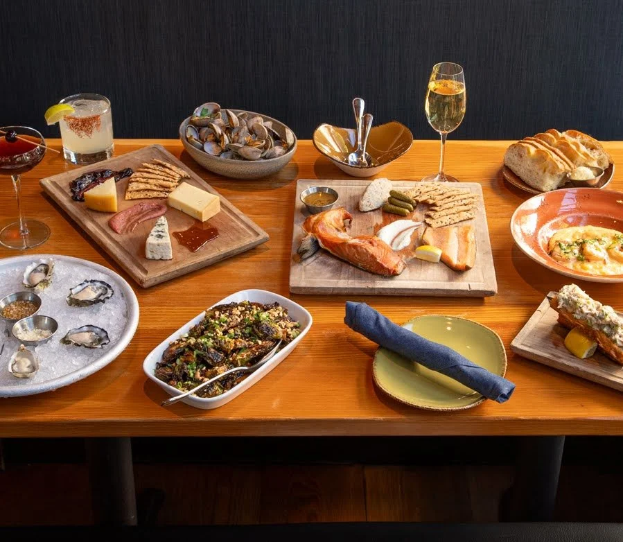 A variety of seafood, cheeses, and appetizers on a wooden table, including clams on ice, a seafood dish, bread, and drinks.