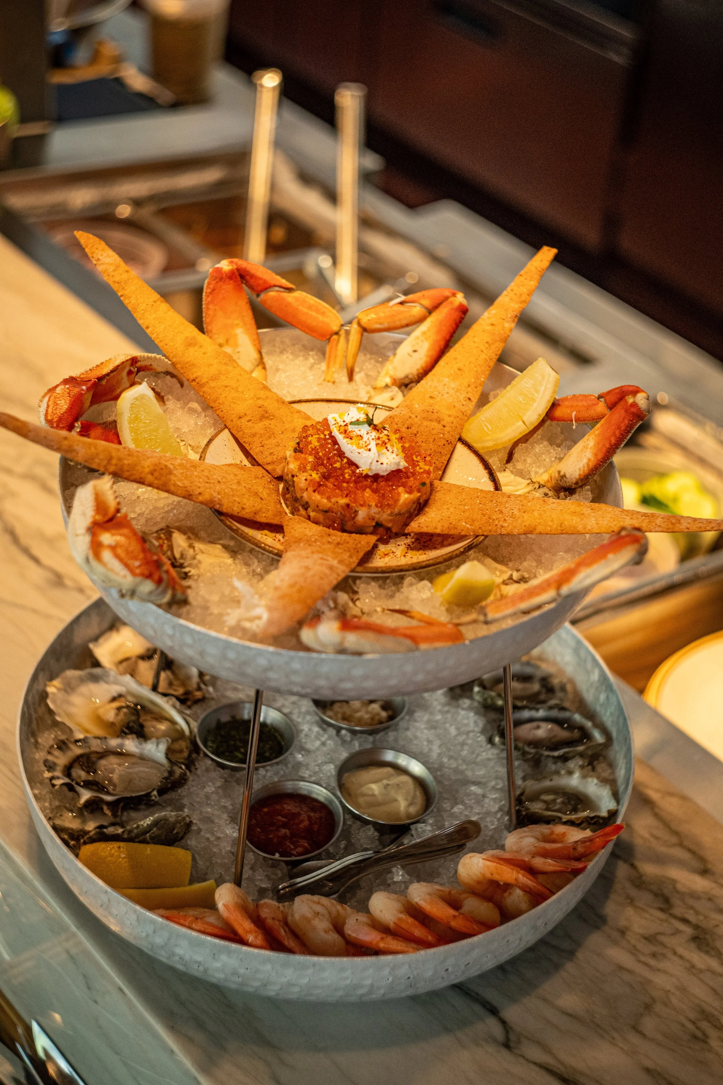 A two-tier seafood platter with shrimp, crab legs, lemon wedges, and various dipping sauces on a bed of ice