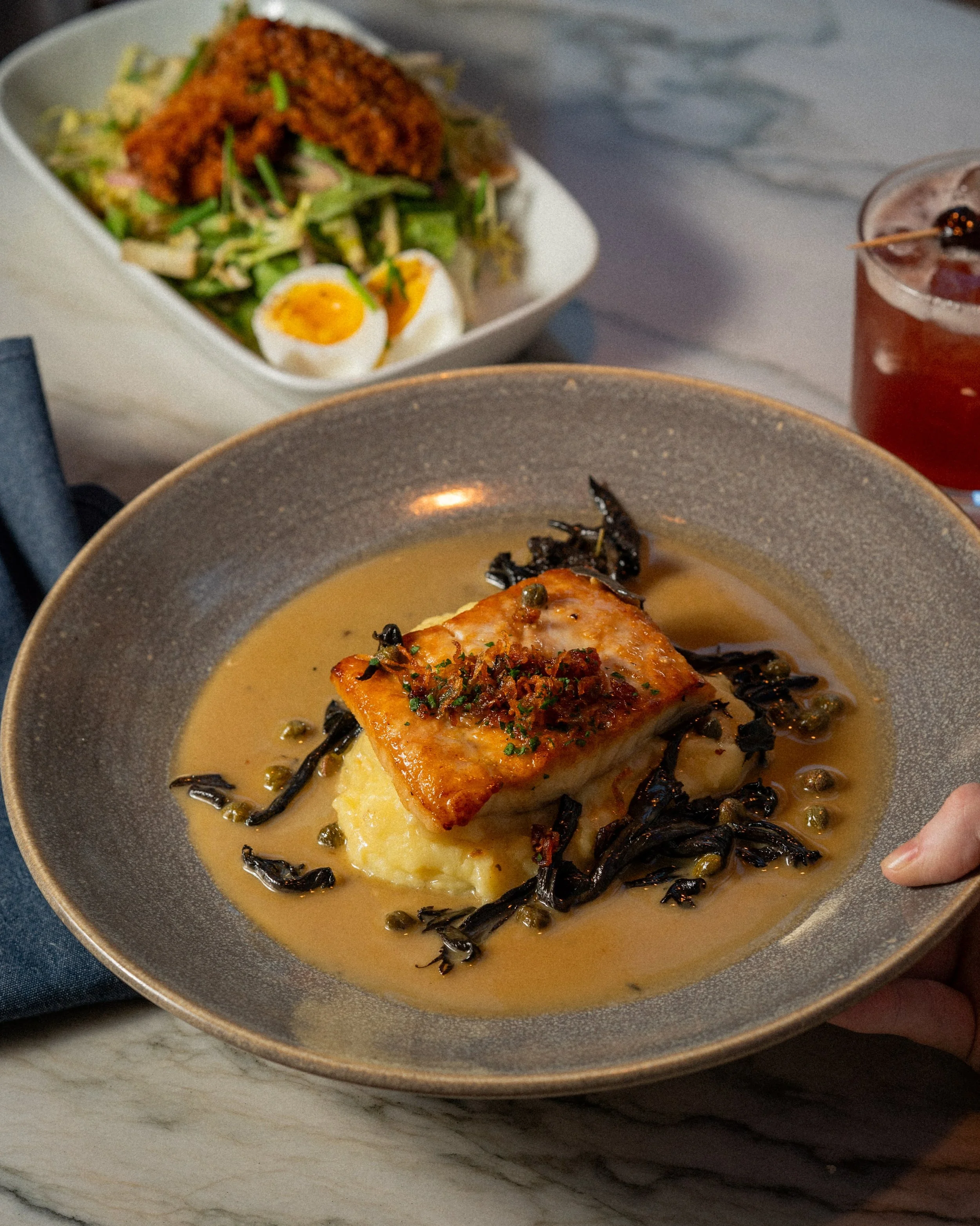 A plated dish with a piece of cooked fish topped with herbs and sauce, surrounded by black pasta in a creamy sauce.