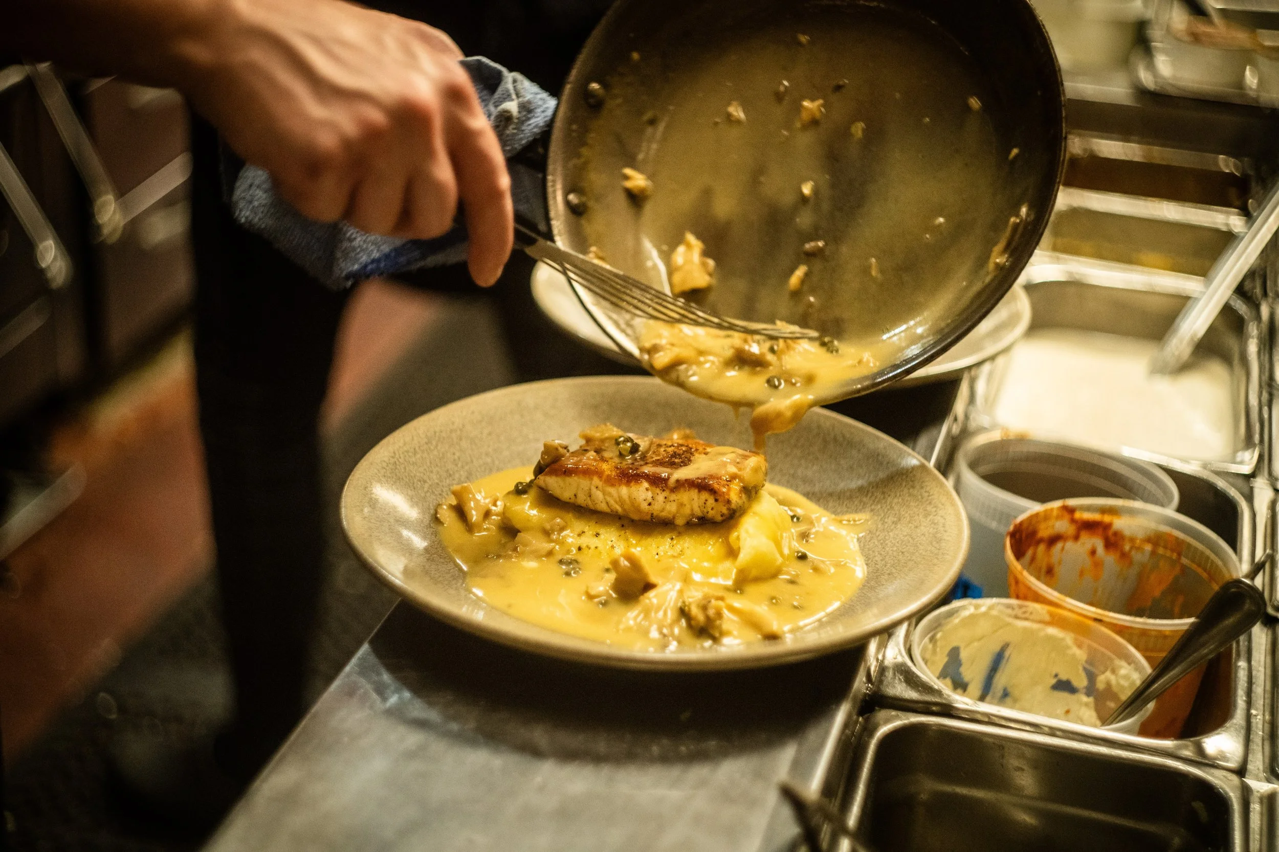 A person pouring a creamy gravy with mushrooms over a plated main dish in a kitchen setting.