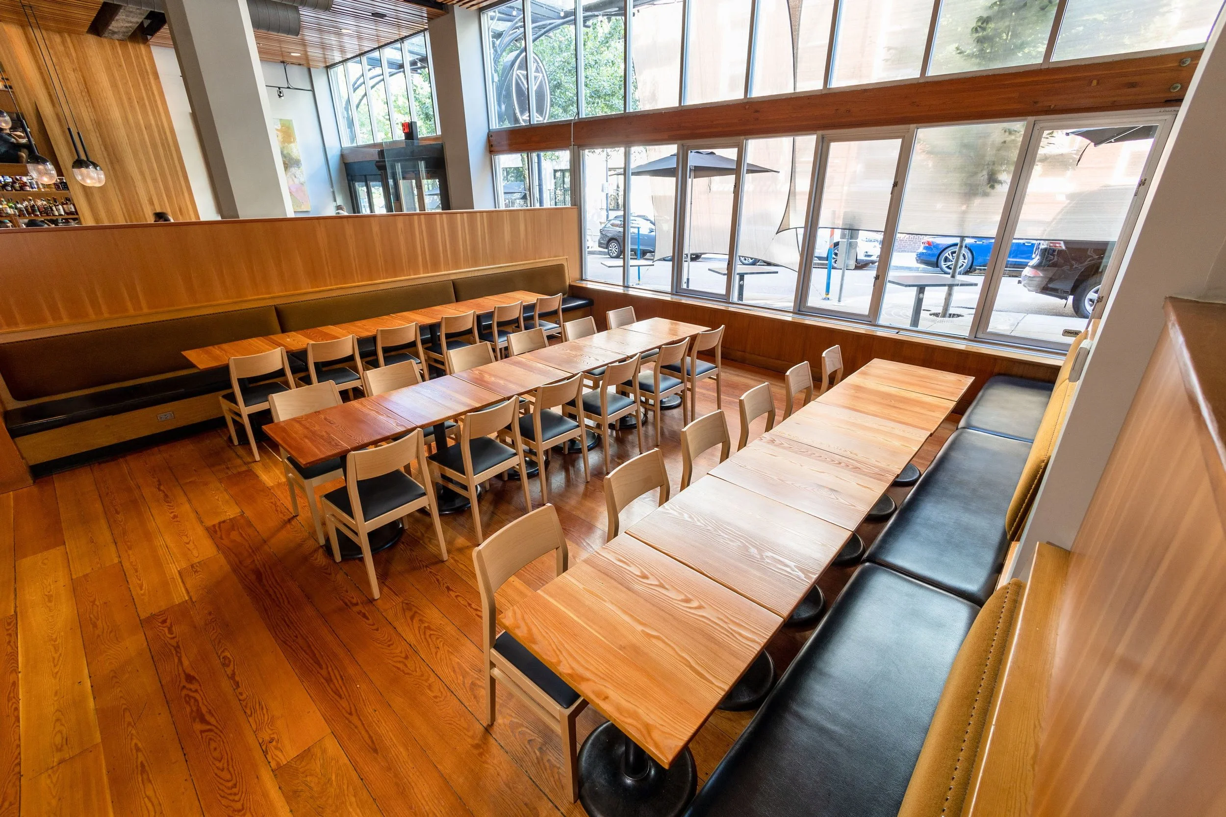 Interior of a modern restaurant or cafe with wooden tables and chairs, large windows, and a wood-paneled ceiling.