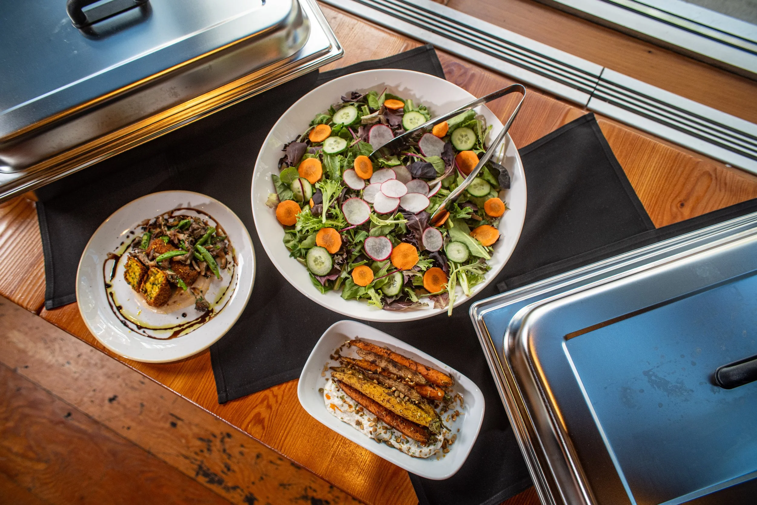 A buffet table with a large bowl of mixed salad containing cucumbers, radishes, and carrots, and smaller dishes with grilled vegetables, a meat dish with sauce, and fried food sticks.