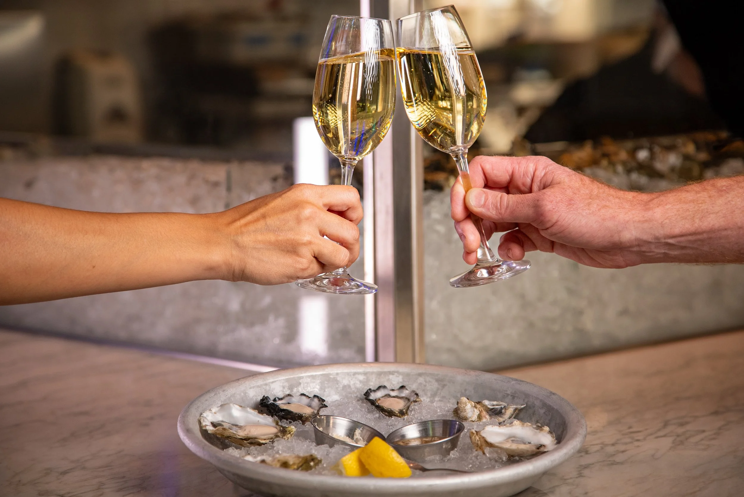 Two people clinking glasses of champagne over a tray of oysters and lemon wedges.