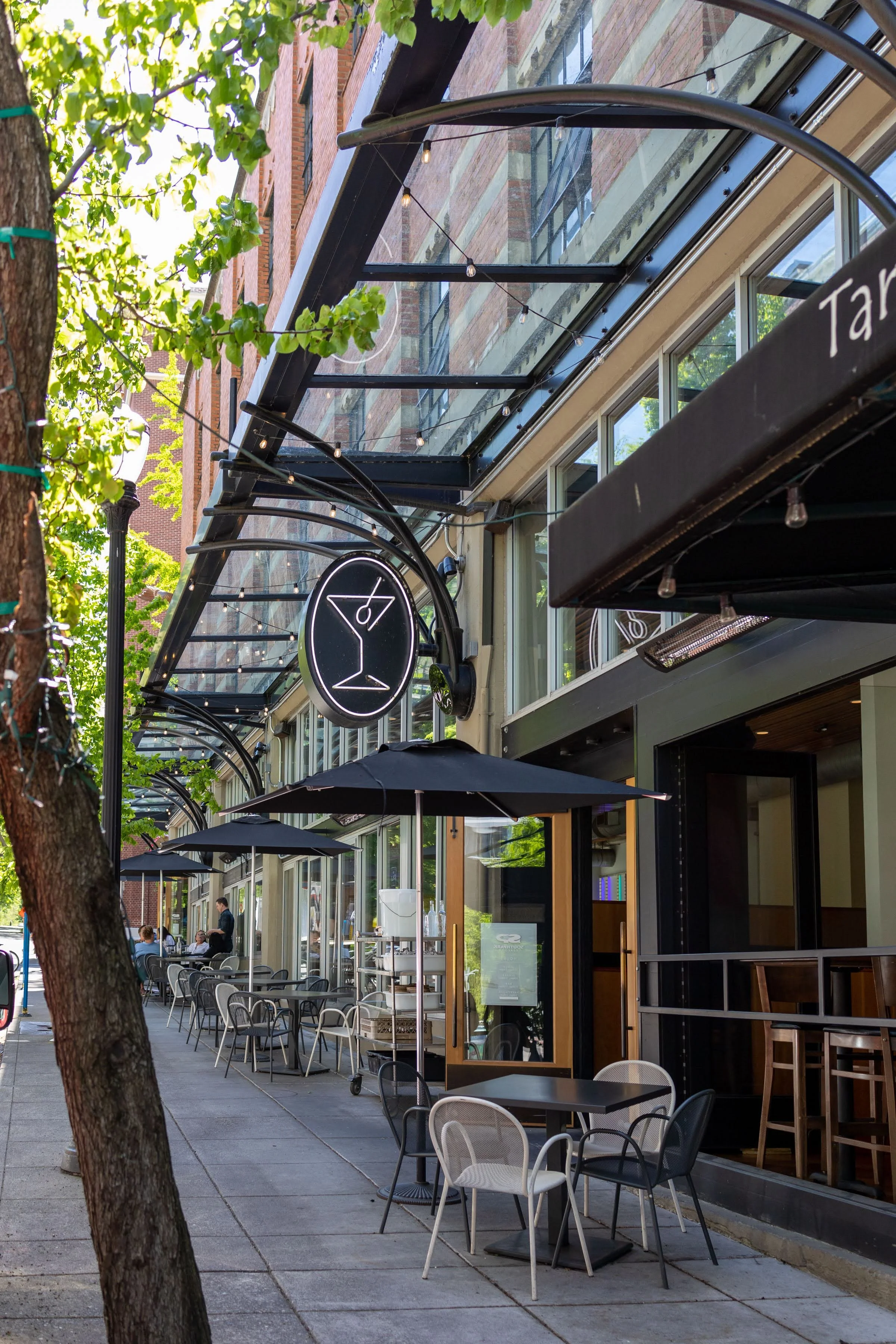 Empty outdoor patio with tables and chairs outside a restaurant or cafe, with black umbrellas and string lights, a tree in the foreground, and a brick building with large windows and a glass canopy.