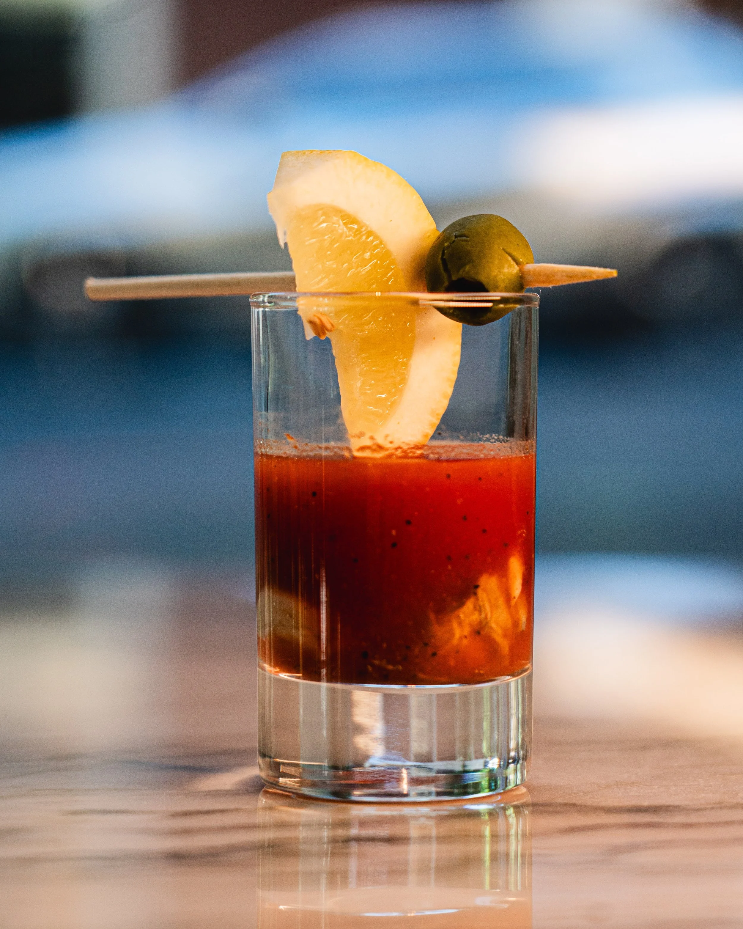 A close-up of a layered cocktail in a clear glass with a skewer holding a green olive, a slice of lemon, and a slice of orange, placed on a wooden surface with a blurred background.
