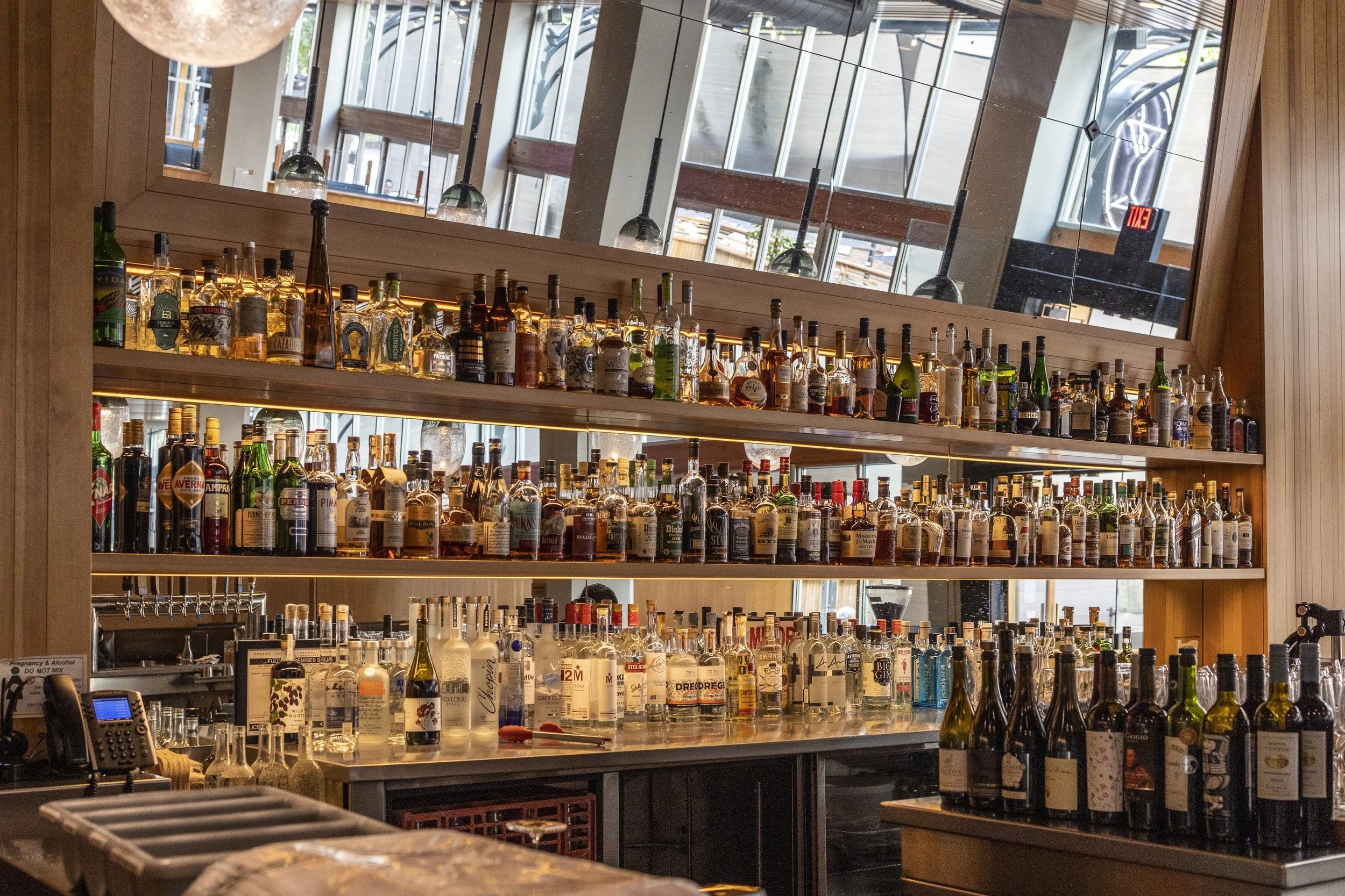 Bar with shelves of various liquor bottles against a large window with reflections and an EXIT sign in the background.
