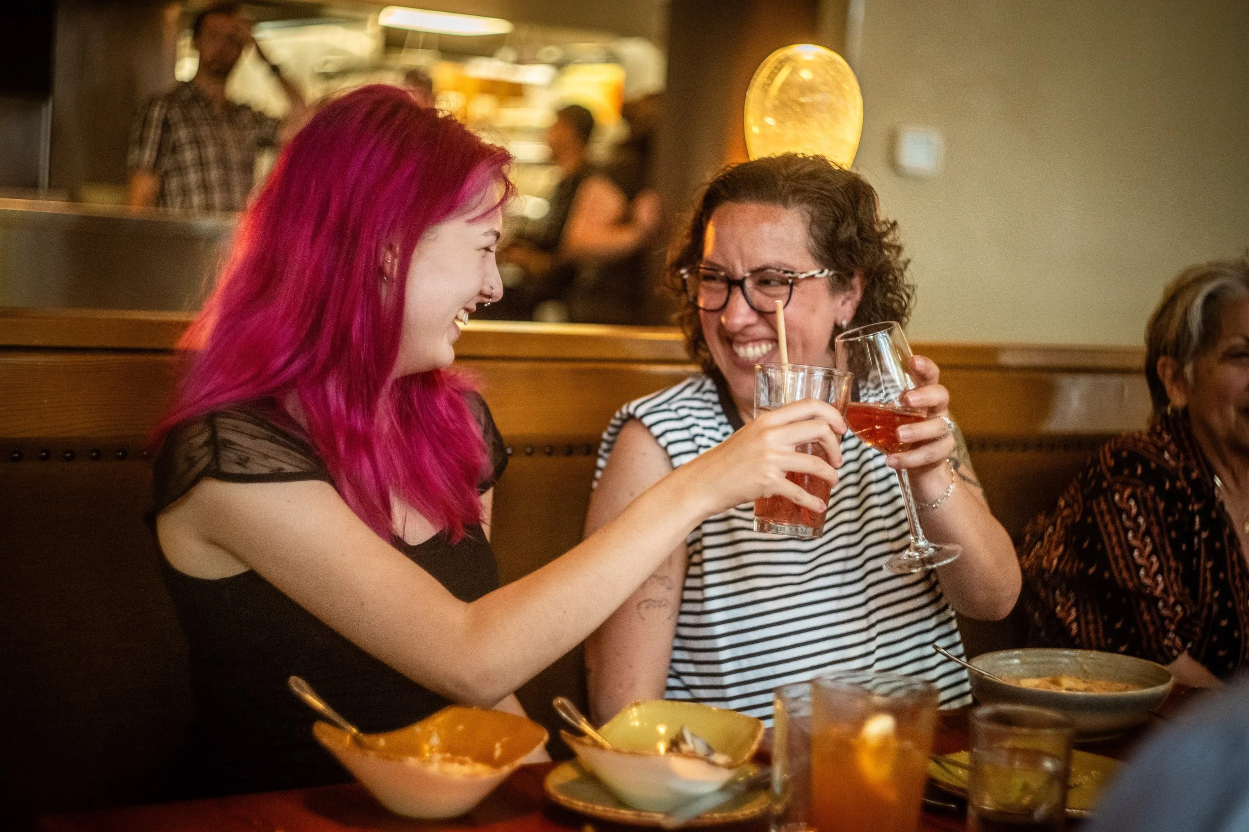 Two women laughing and clinking glasses in a restaurant. One has pink hair and the other has short, curly brown hair and glasses. There are food bowls and drinks on the table.