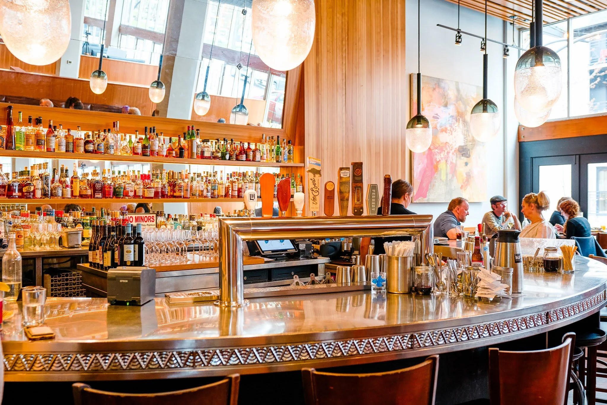 Interior of a modern restaurant or bar with a curved wooden bar counter, bottles of alcohol on shelves, multiple hanging pendant lights, and diners seated at tables.