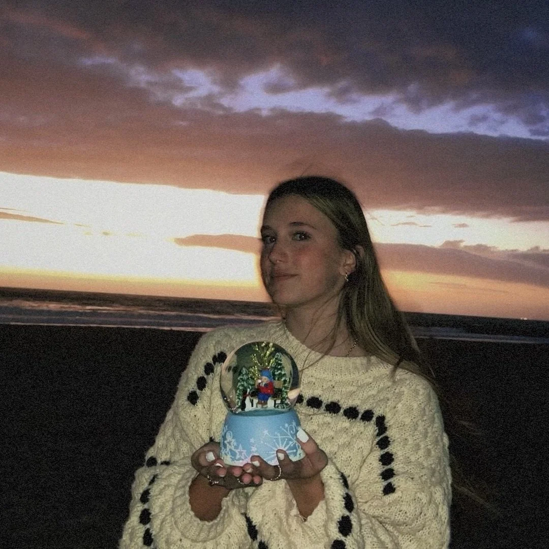 A woman holding a snow globe on a beach at sunset, with the ocean and colorful sky in the background.