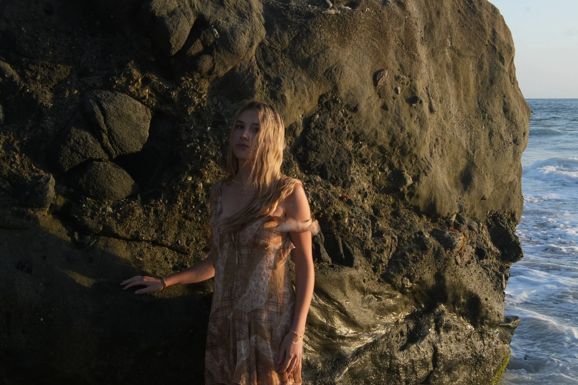 A young woman with long wavy hair sitting by rocky coastal shoreline with ocean waves in background.