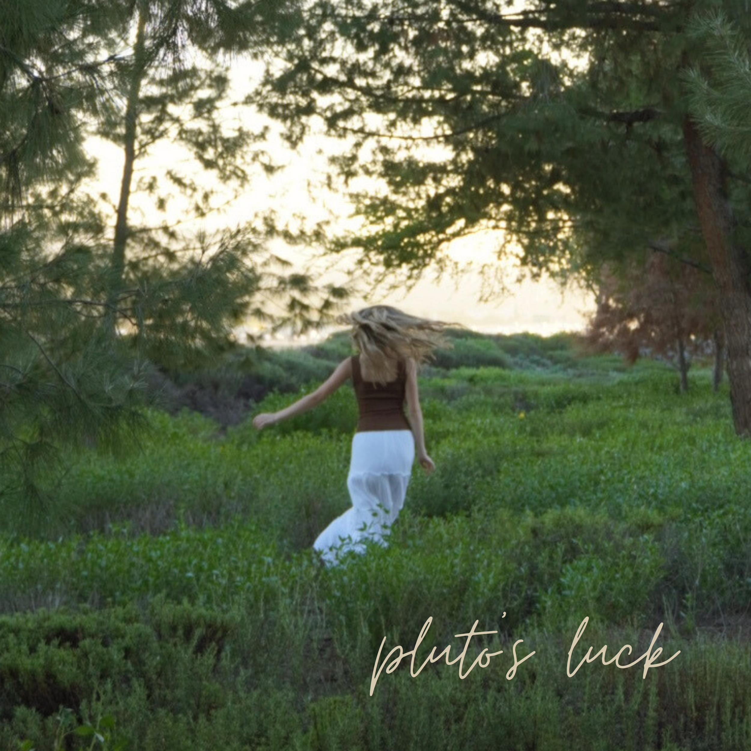 Woman with blonde hair wearing a brown top and white pants stands in a grassy area among tall green trees, with sunlight filtering through the branches.