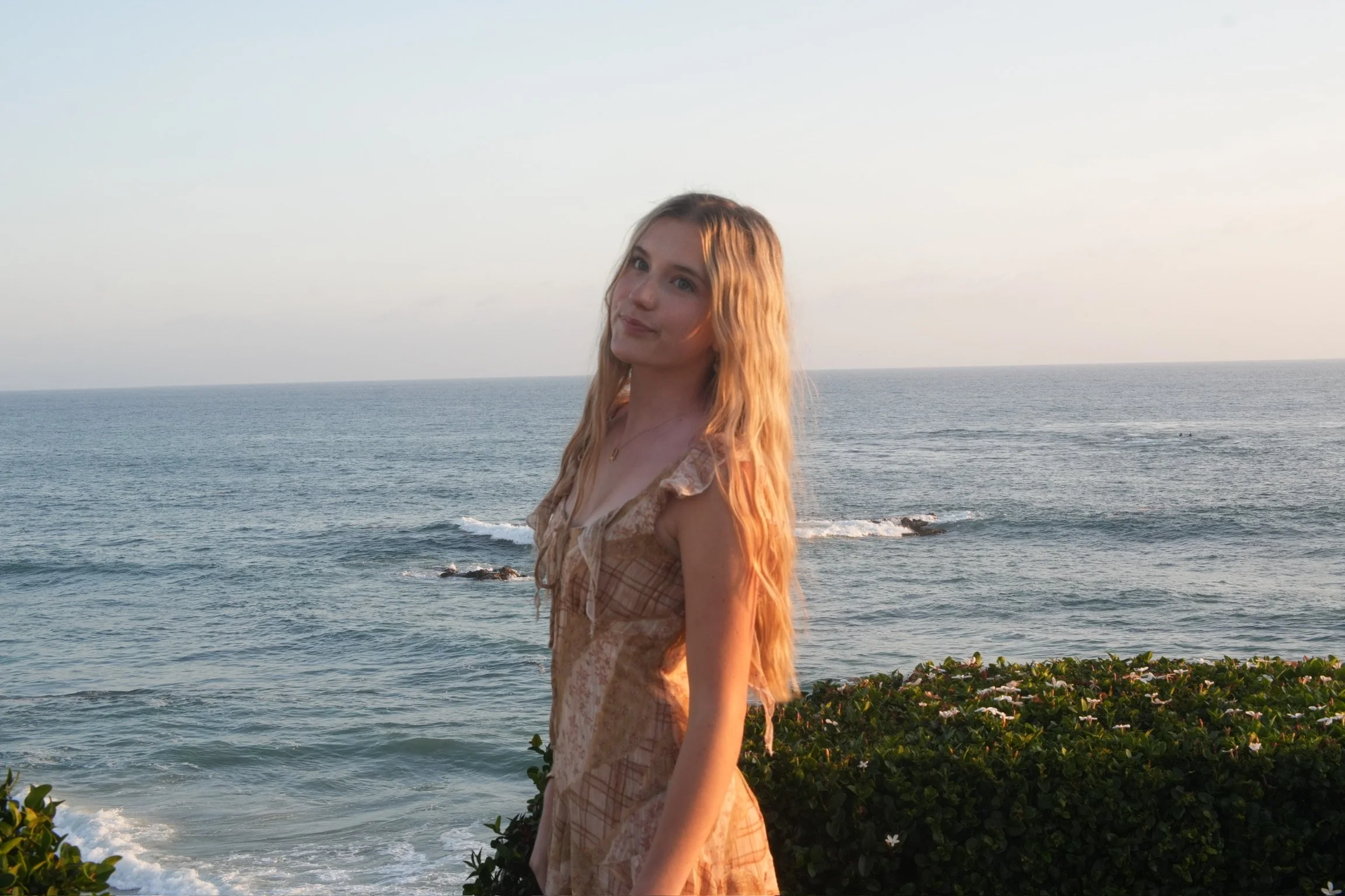 A young woman with long, wavy, blonde hair standing by the ocean at sunset, wearing a light brown patterned dress, with green bushes in the foreground.
