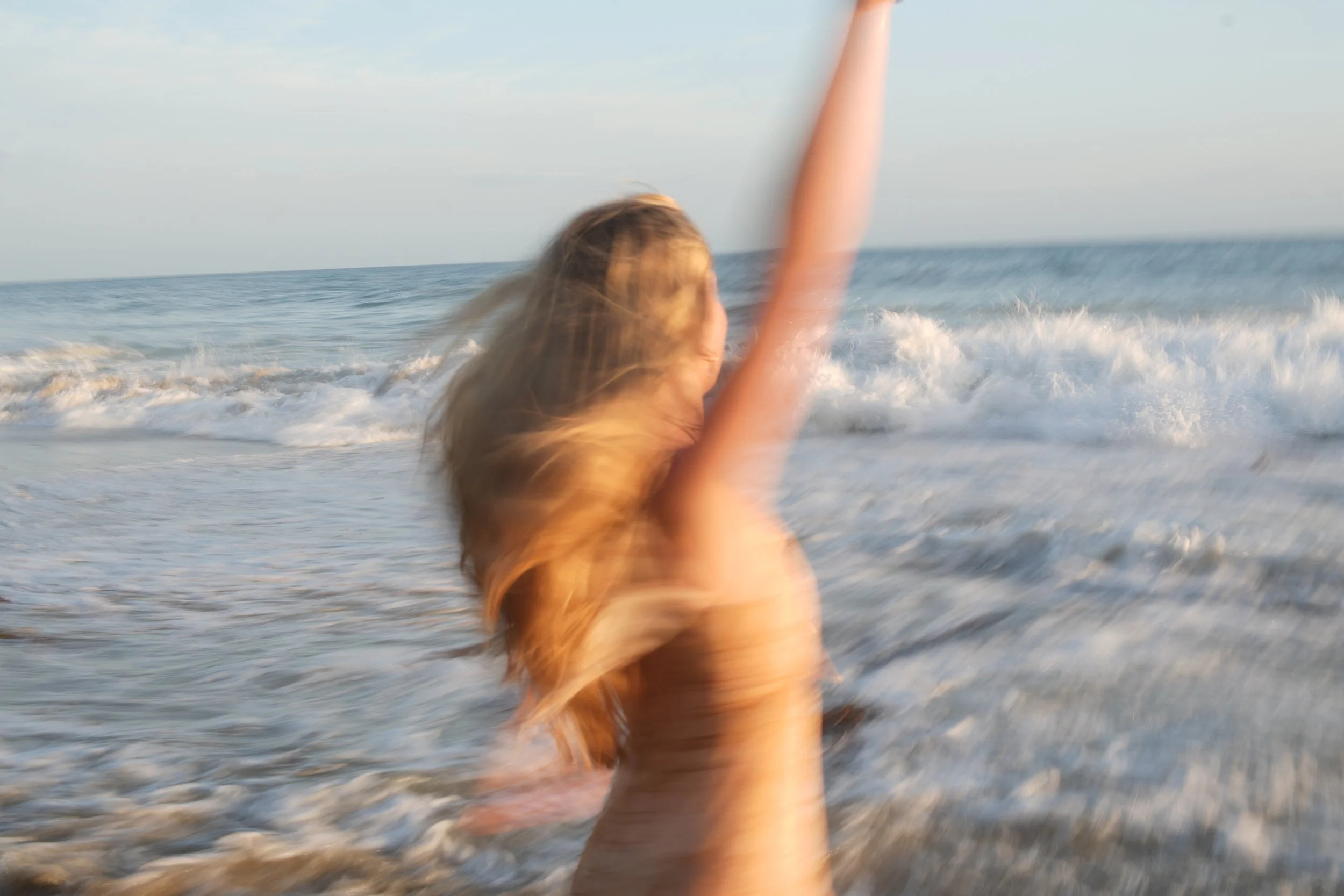 A person with long hair in a tan shirt is at the beach, raising an arm. The ocean waves are in the background, and the scene is illuminated by natural sunlight.