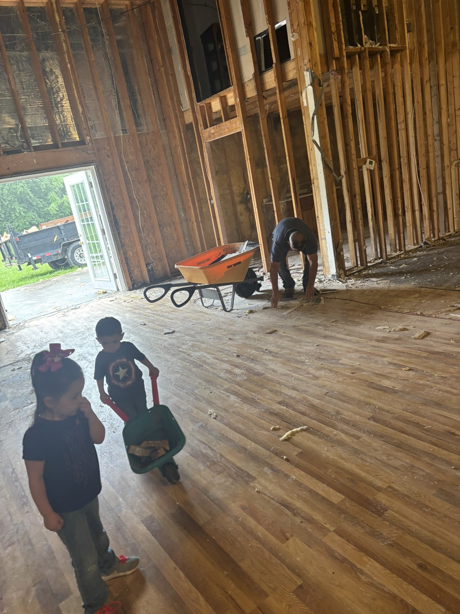 A person is using a tool on a wooden floor inside a house under renovation. There are children playing in the foreground, and a wheelbarrow with construction debris is nearby. The walls are exposed with visible wooden studs, and there is an open gara